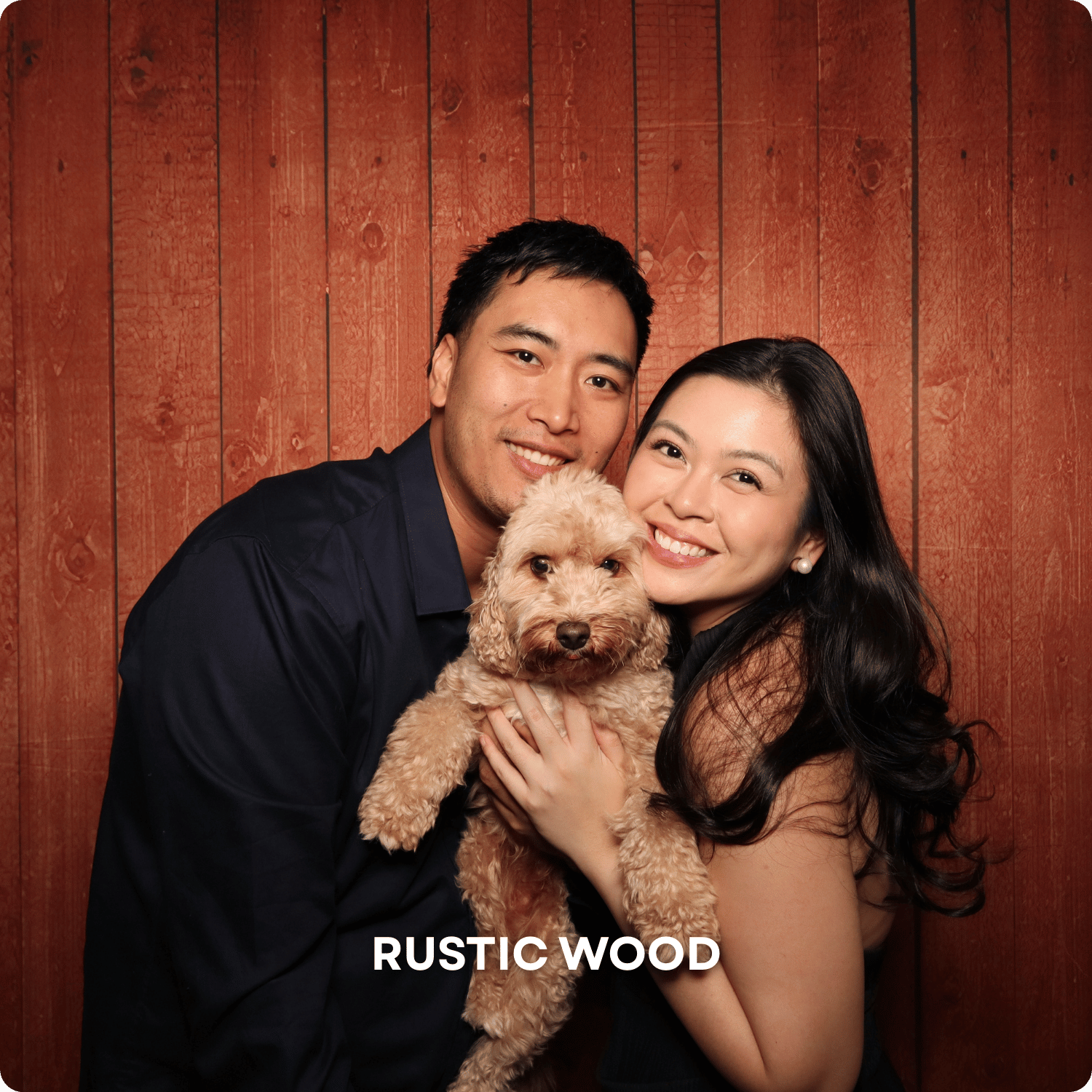 A smiling couple with light skin holding a light brown curly-haired dog in front of a rustic wooden wall.