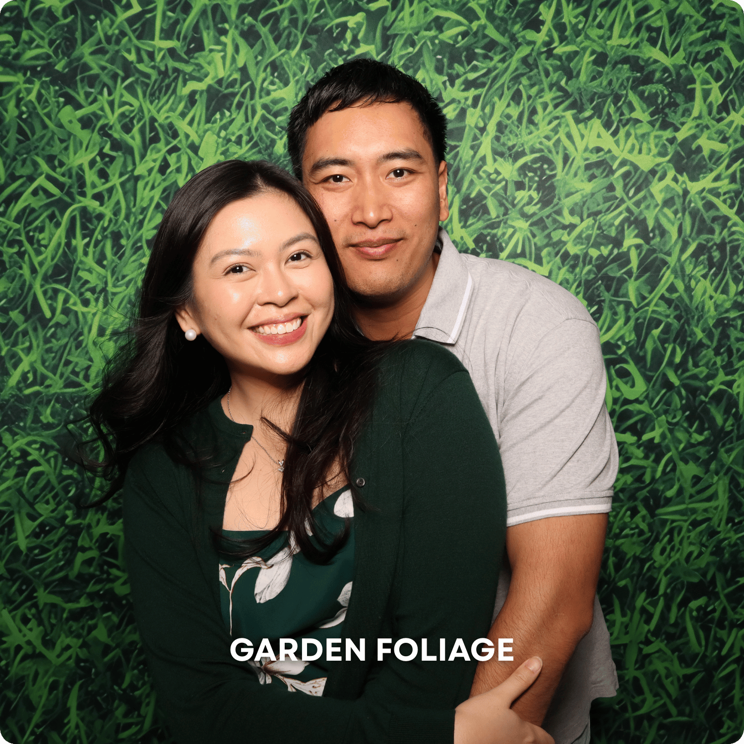 A smiling Asian woman with long dark hair, wearing a green dress and pearl earrings, is embracing a smiling Asian man with short dark hair, wearing a light gray polo shirt. They are standing in front of a green garden foliage backdrop with the text "GARDEN FOLIAGE" at the bottom.