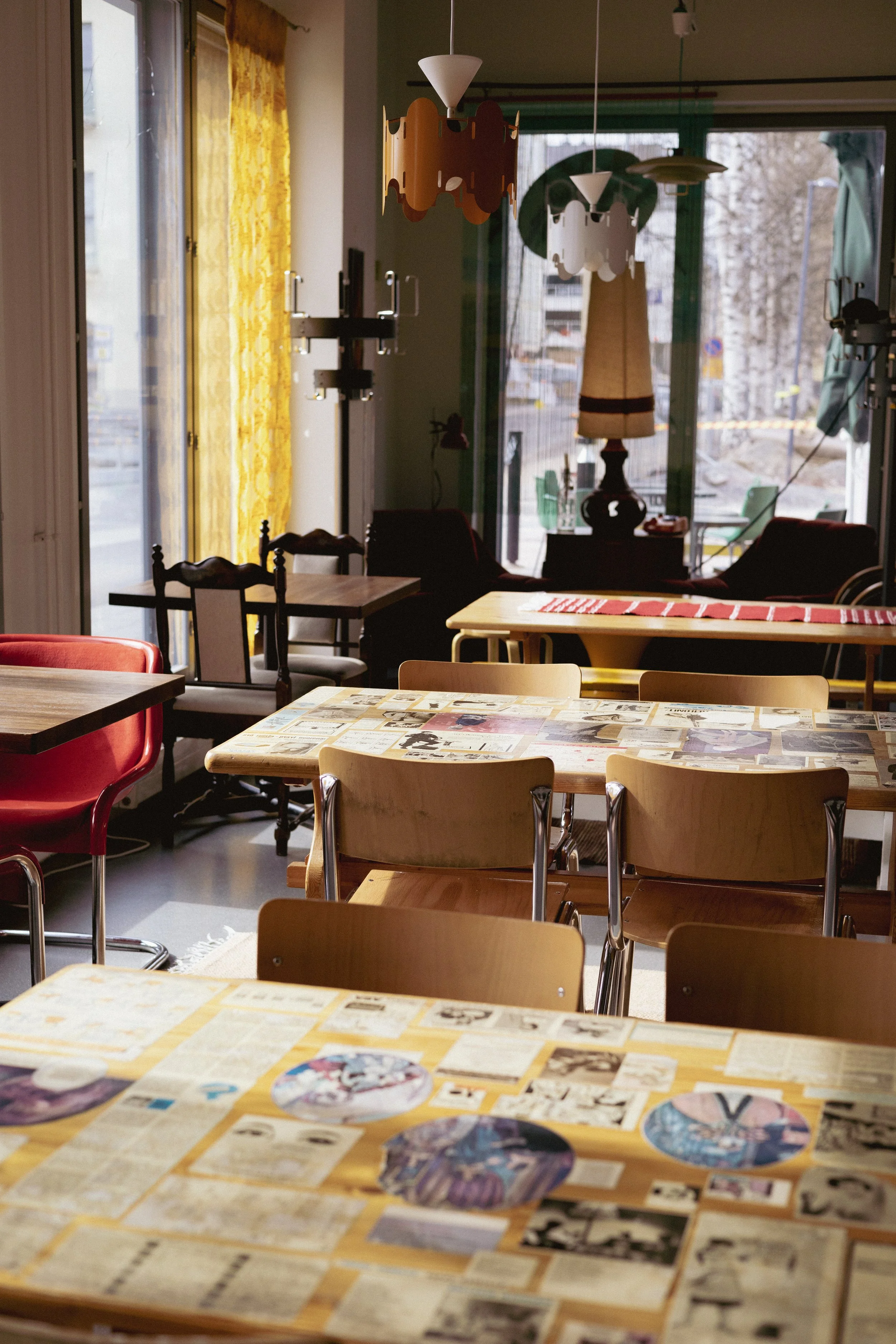 Kaksi pöytää sisätiloissa, kaksi pöytää ulkona näkyvissä, sisältä näkee ikkunoista ja oville ulos. Sisätiloissa on erilaisia tuoleja ja joukossa on pöytä, jonka pinnalla on erilaisia lehtiä tai kuvia.