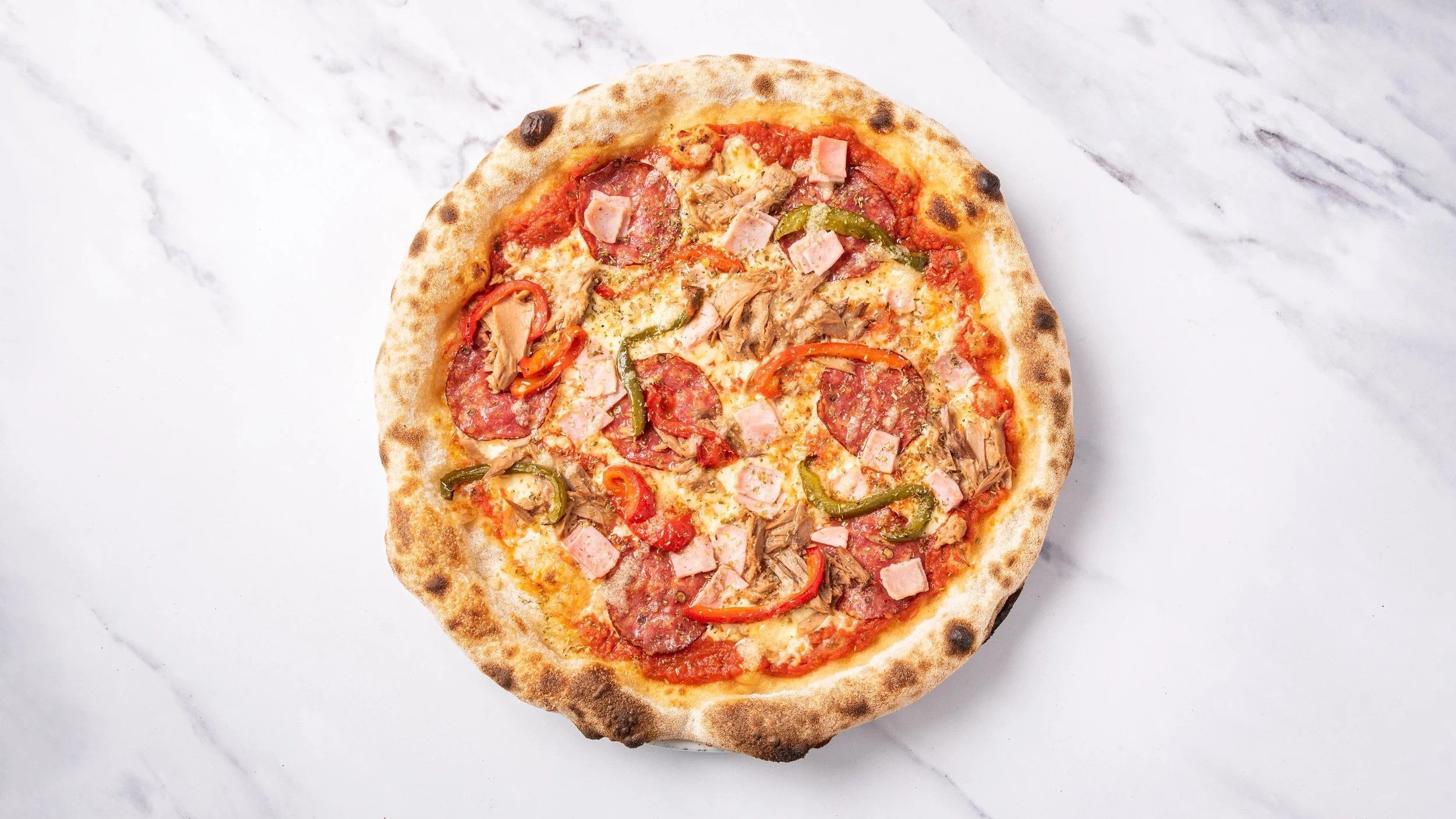 Kuvassa pizzaa, jossa on pepperoniä, kinkkua, tonnikalaa ja vihanneksia, kuten paprikaa ja punasipulia, tarjoiltuna valkoisella marmoripöydällä.