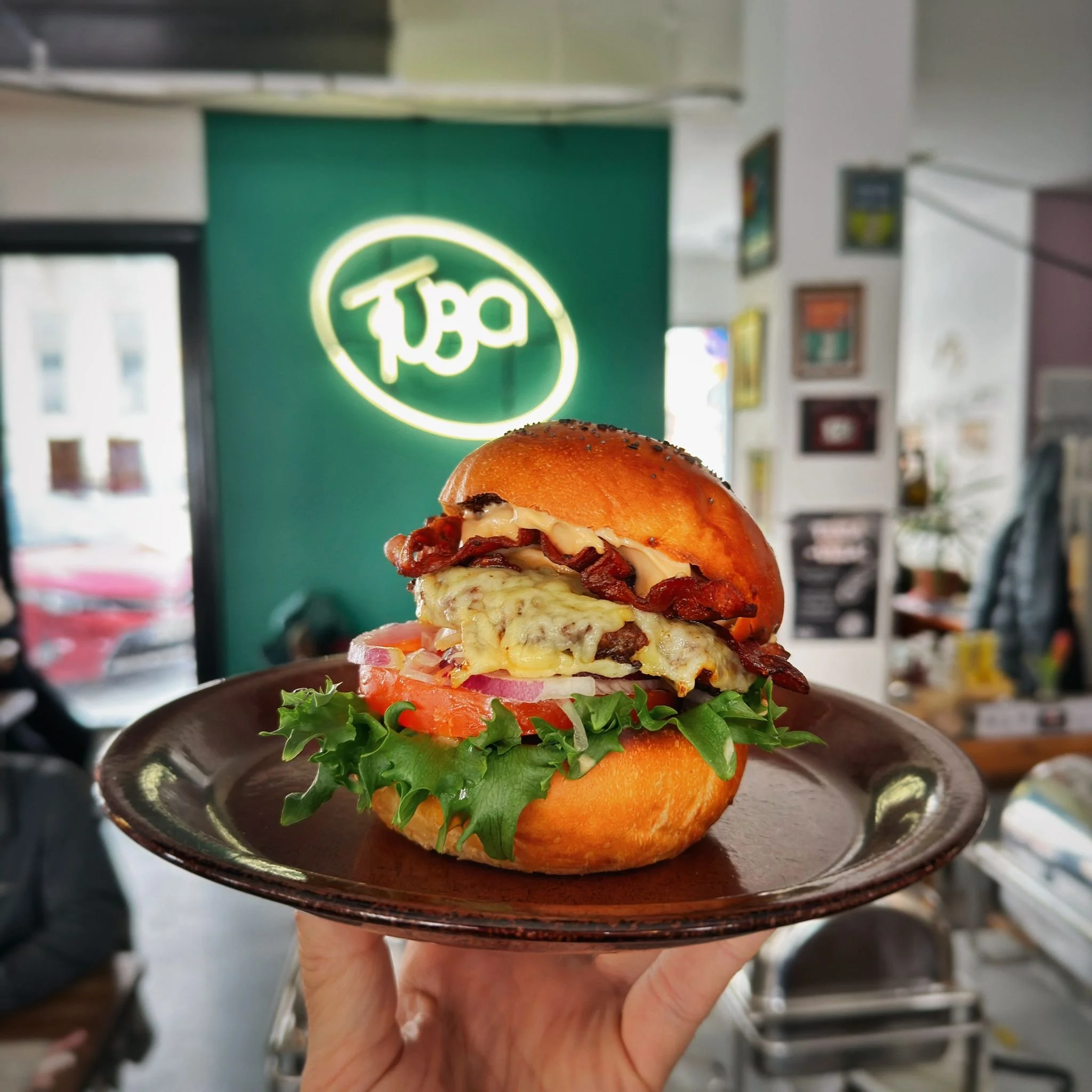 Henkilö pitää lautasella hampurilaista, jossa on leipää, kasviruokaa, juustoa, lihaa, tomaattia, sipulia ja salaattia. Taustalla neonvalomainos ja sisätilojen ravintolaympäristö.