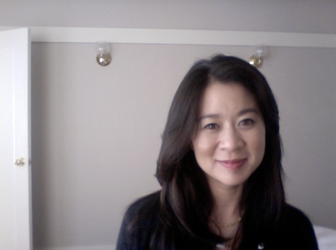Cindy Kay with long dark hair smiling at the camera in a room with a beige wall and some small sockets or fixtures on the ceiling.
