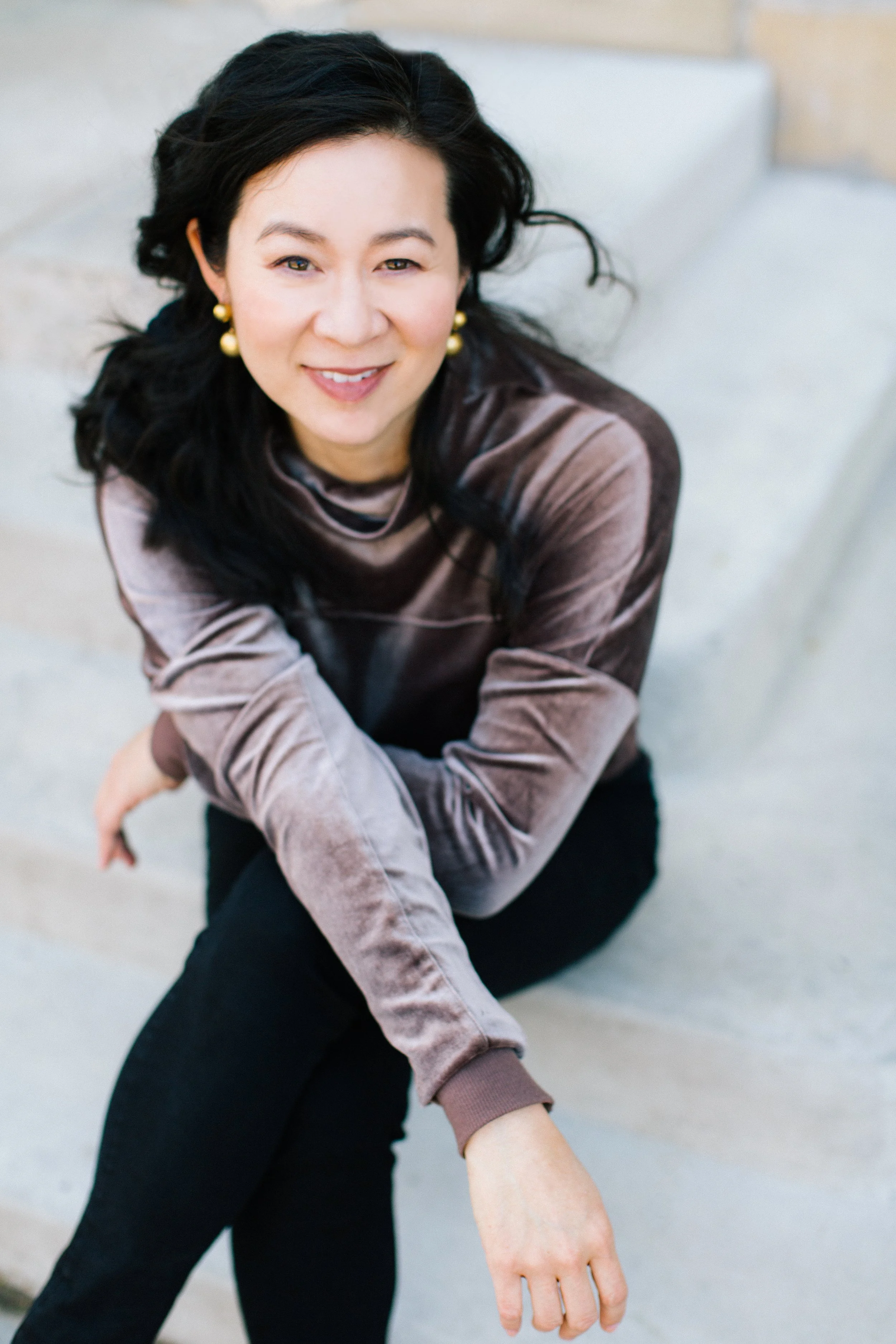 Cindy Kay sitting on steps, smiling, wearing a velvet top and gold earrings.