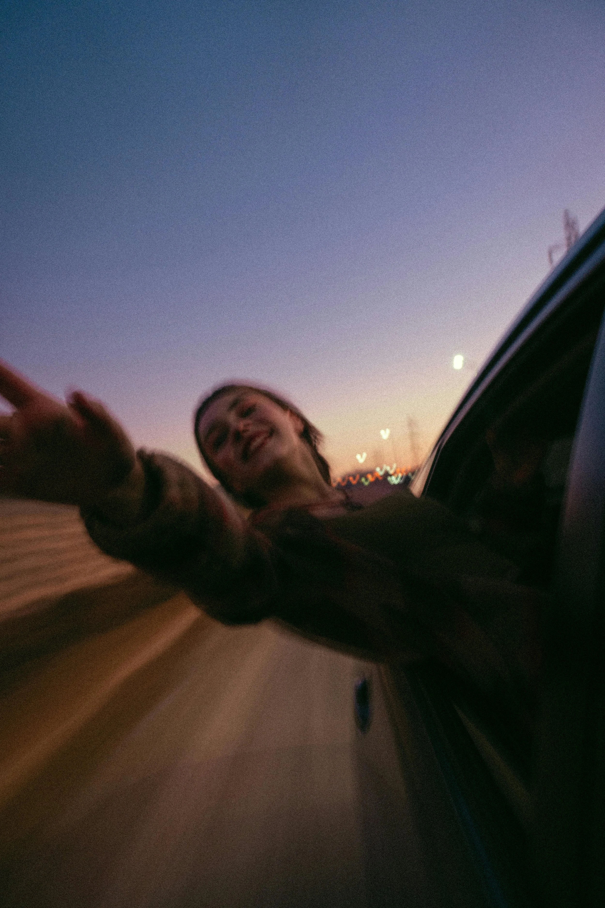 A woman with a happy expression leans out of a car window during sunset, extending her hand.