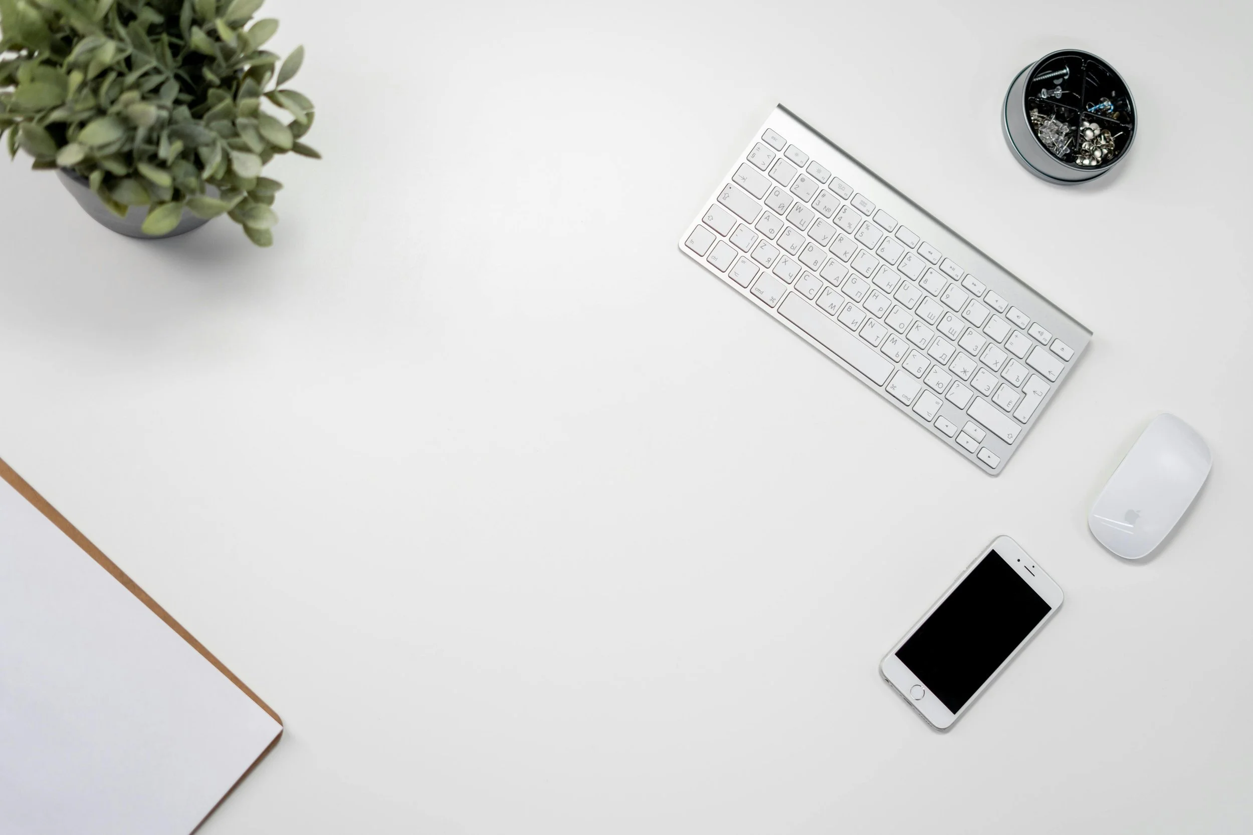 White desk with a potted green plant, a white keyboard, a white computer mouse, a smartphone, and a round container filled with various small jewelry pieces.