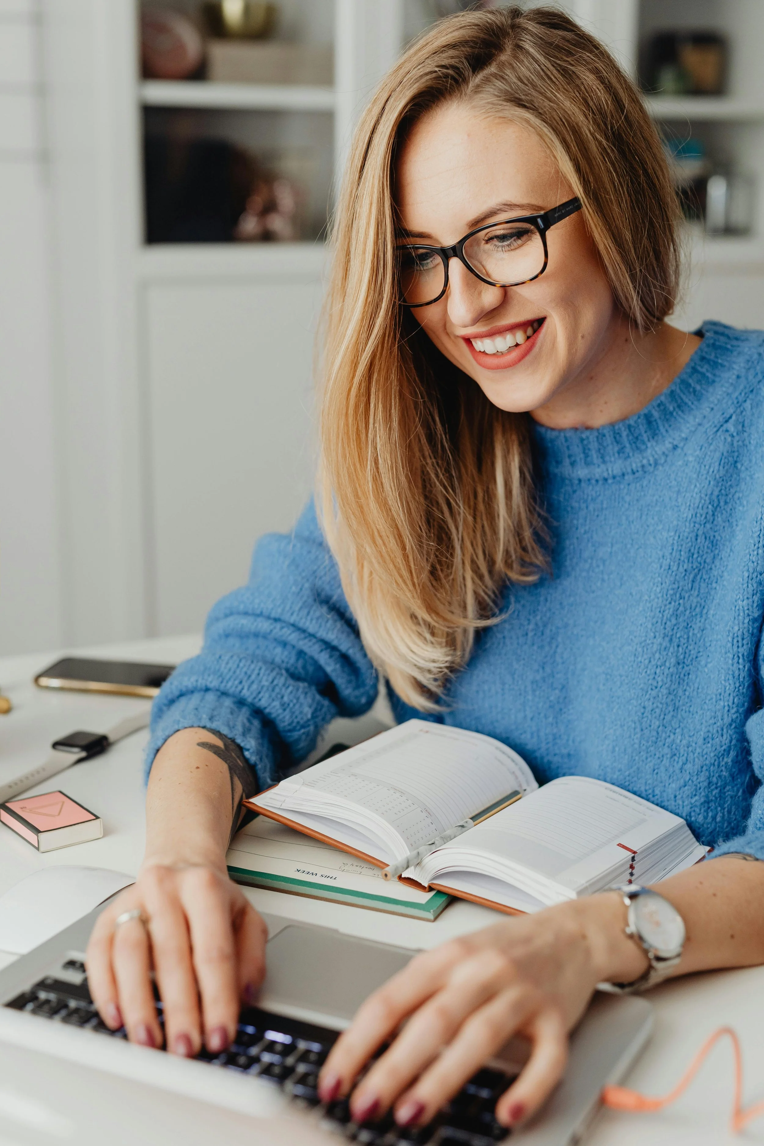 A woman with blonde hair and glasses smiling while working on a laptop at a desk with notebooks and a smartphone.