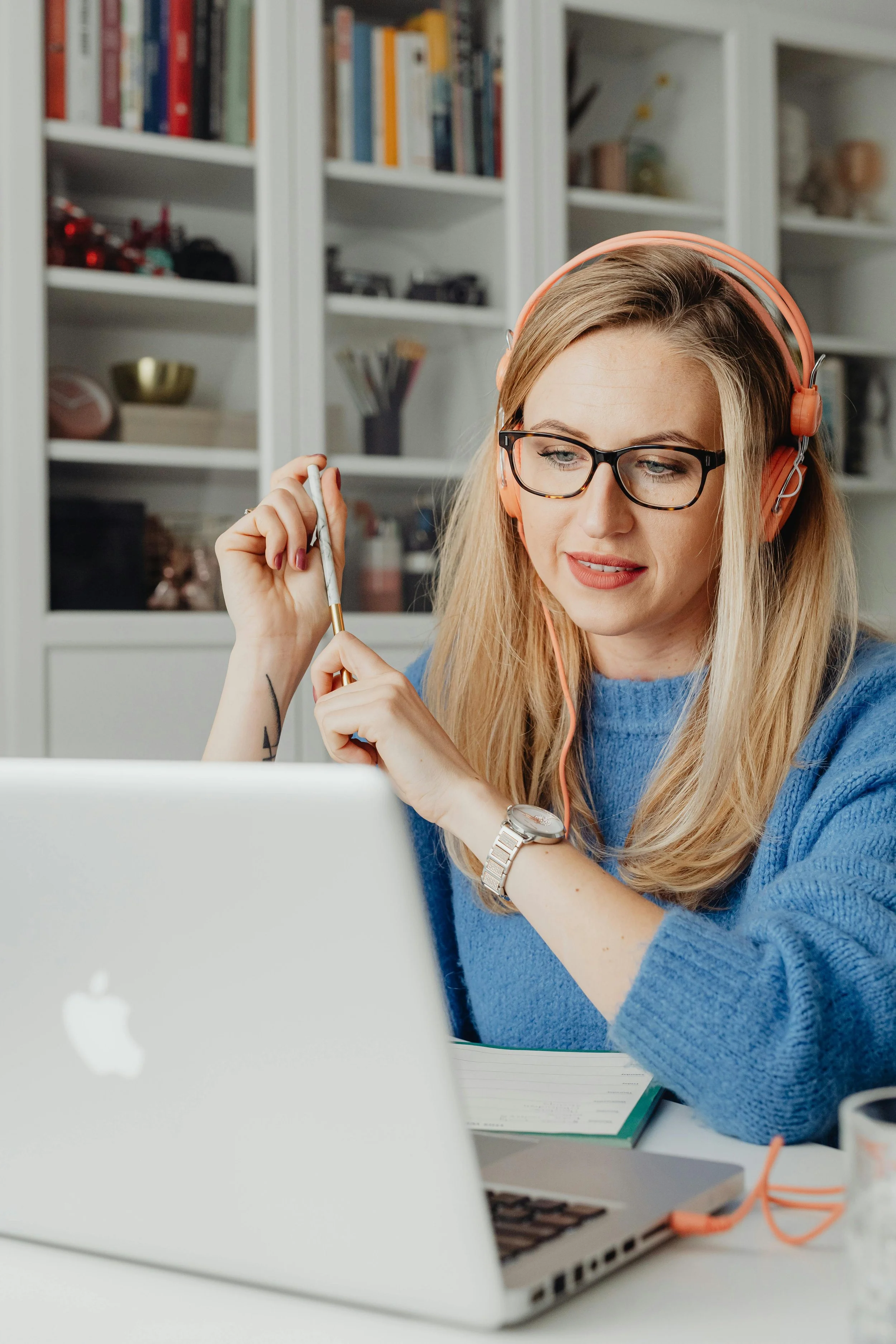 A woman wearing glasses and a blue sweater is sitting at a desk with a laptop, holding a pencil and looking at the screen. She has headphones on and a watch on her wrist.