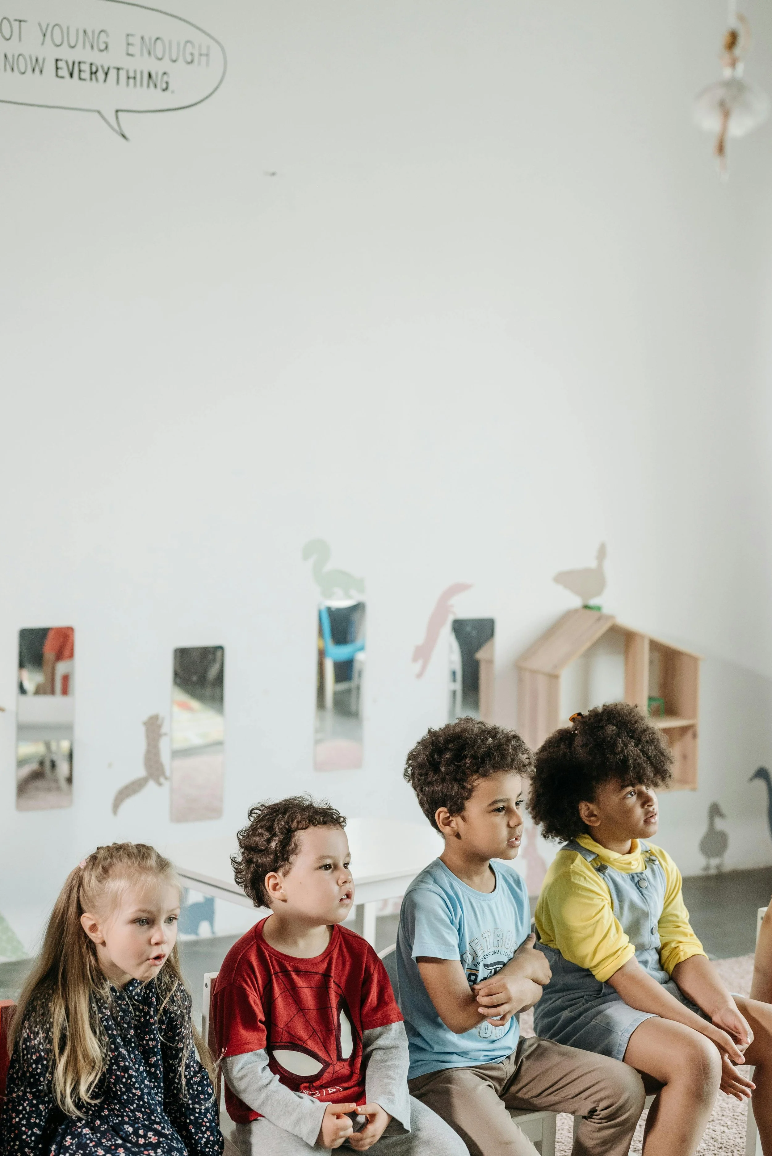Four young children sitting attentively in a classroom with a white wall decorated with animal silhouettes and a quote bubble.