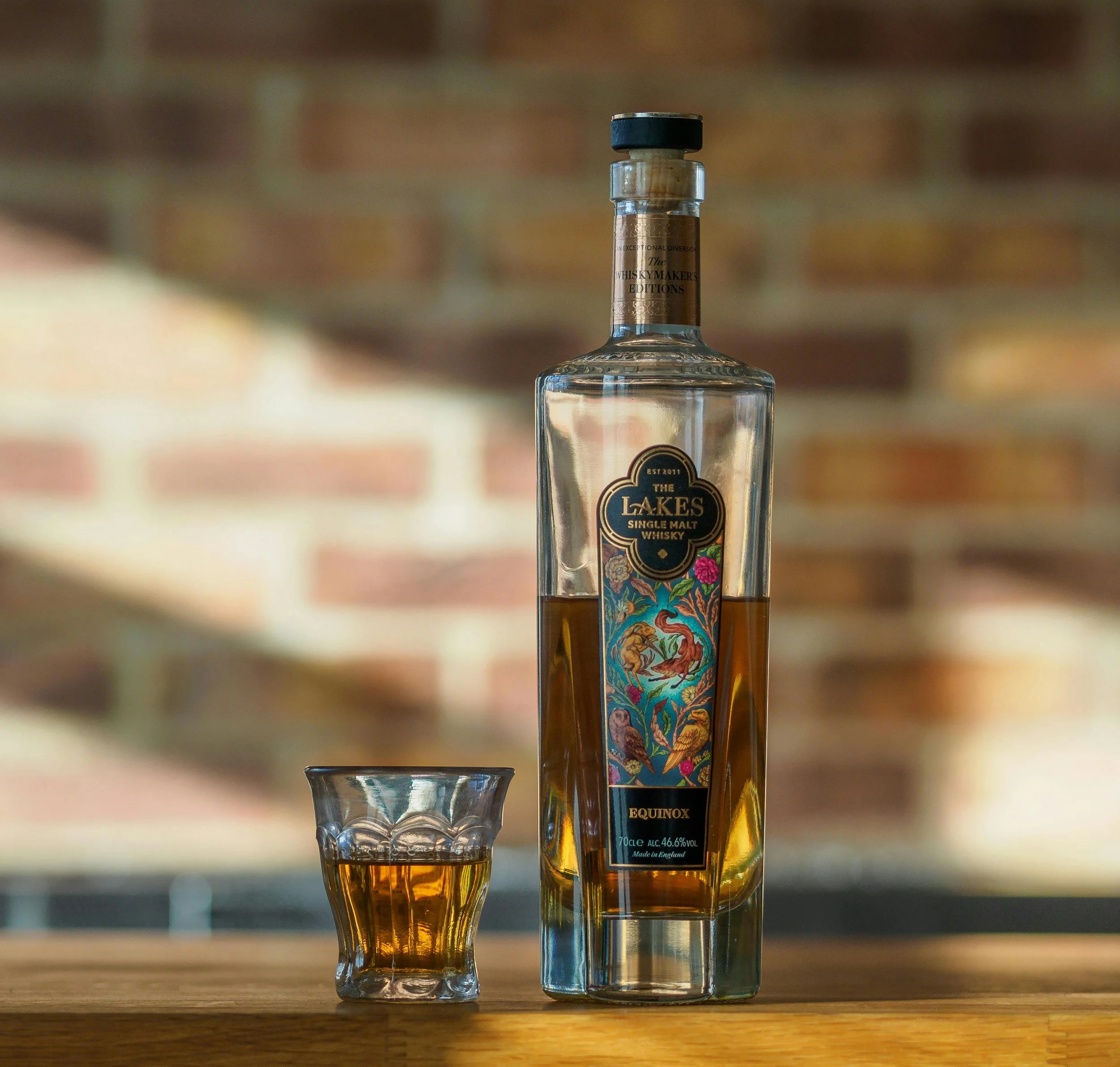 A bottle of The Lakes single malt whisky with a colorful label featuring birds and flowers, placed on a wooden surface next to a glass filled with whisky. The background is blurred with a brick wall.