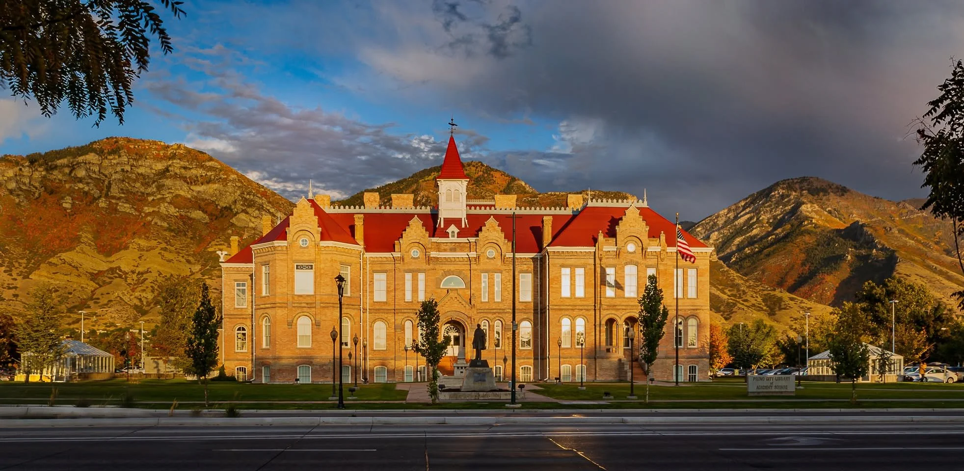 Historic brick courthouse building with red roof, flagpoles, and statue in front, set against mountainous landscape under partly cloudy sky.