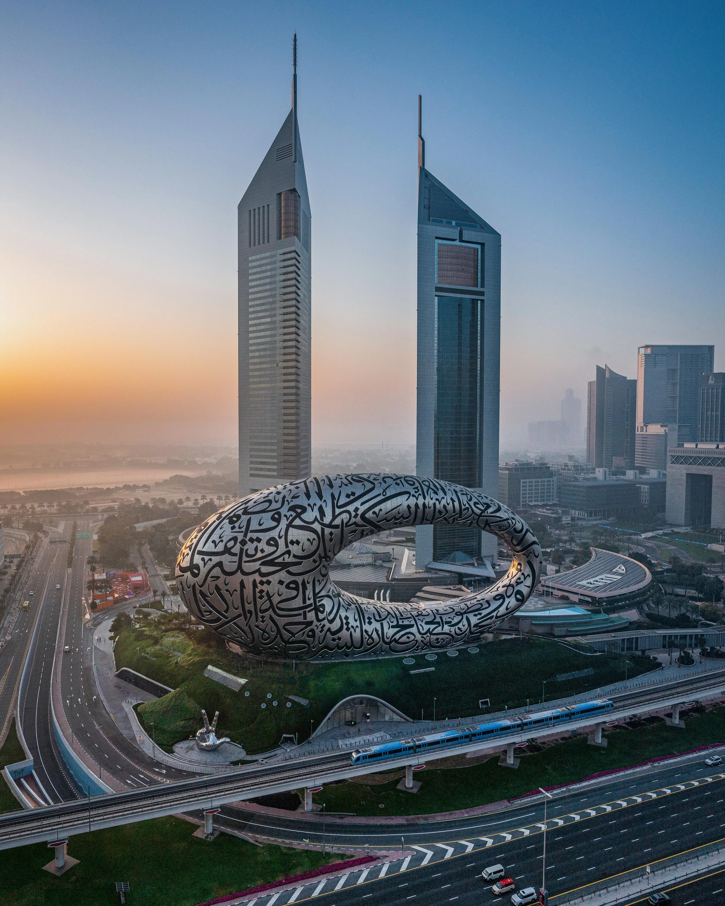 An aerial view of a modern city skyline at sunset featuring two tall skyscrapers and a large, futuristic sculpture with Arabic calligraphy in the foreground.