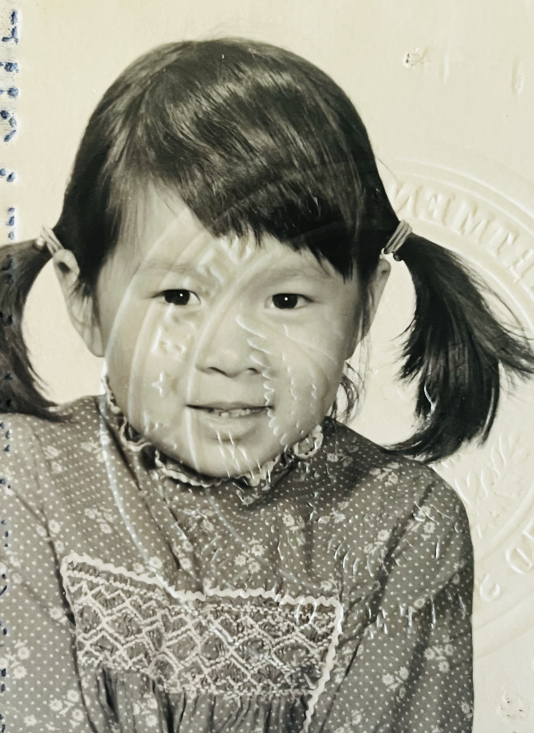 A black and white photo of a young girl with pigtails, wearing a patterned dress and a beaded necklace, sitting on a coin with a profile of Abraham Lincoln, with a neutral wall in the background.