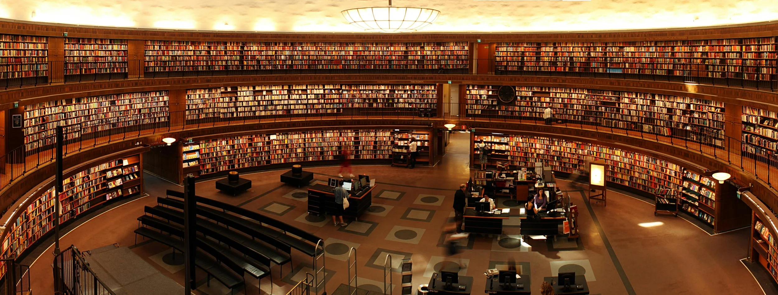 Interior of a large, multi-level library with tall bookshelves made of wood, filled with books. There are tables, computers, and people browsing or working, under warm lighting.