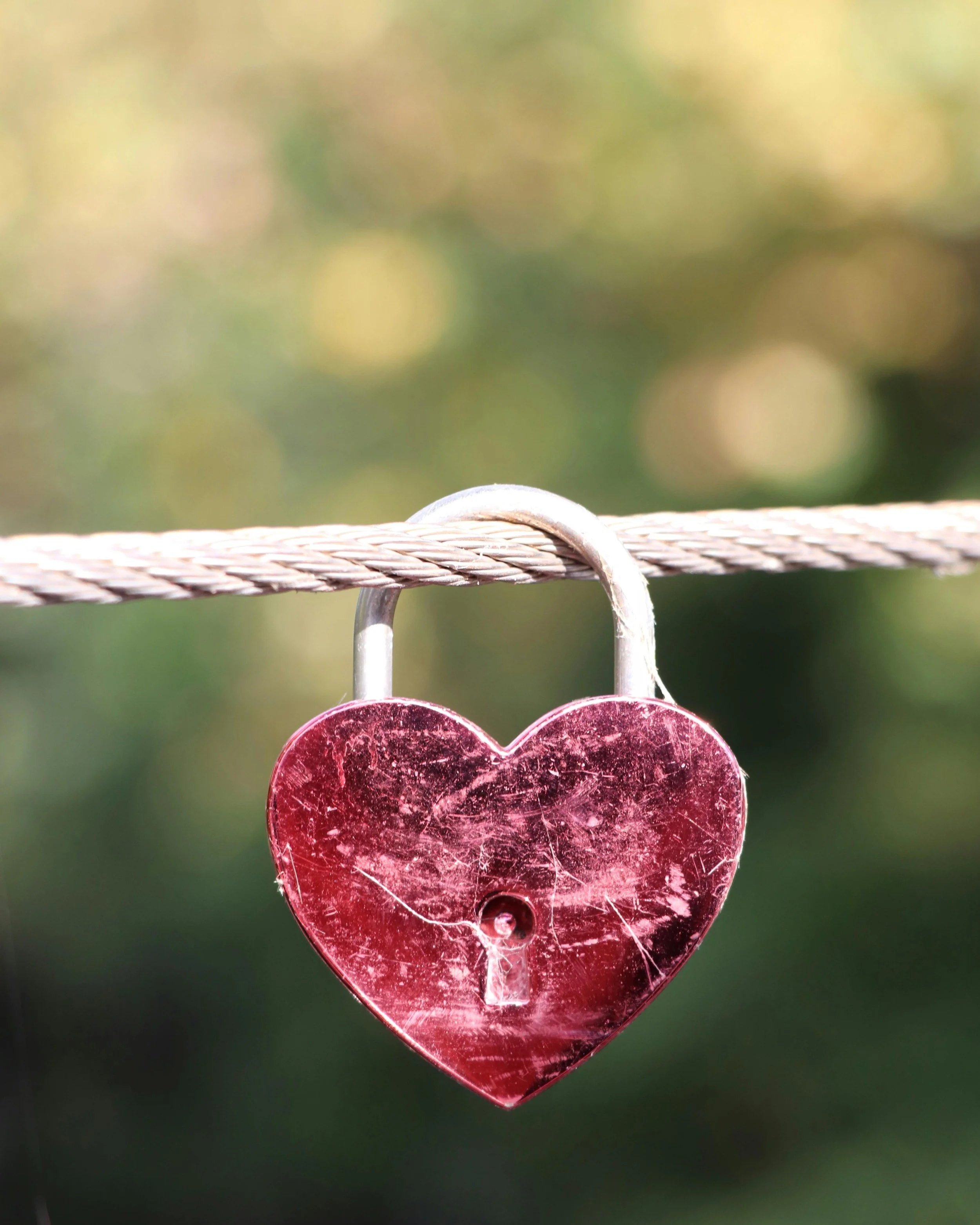A red heart-shaped padlock hanging on a horizontal rope, with a blurred natural background.
