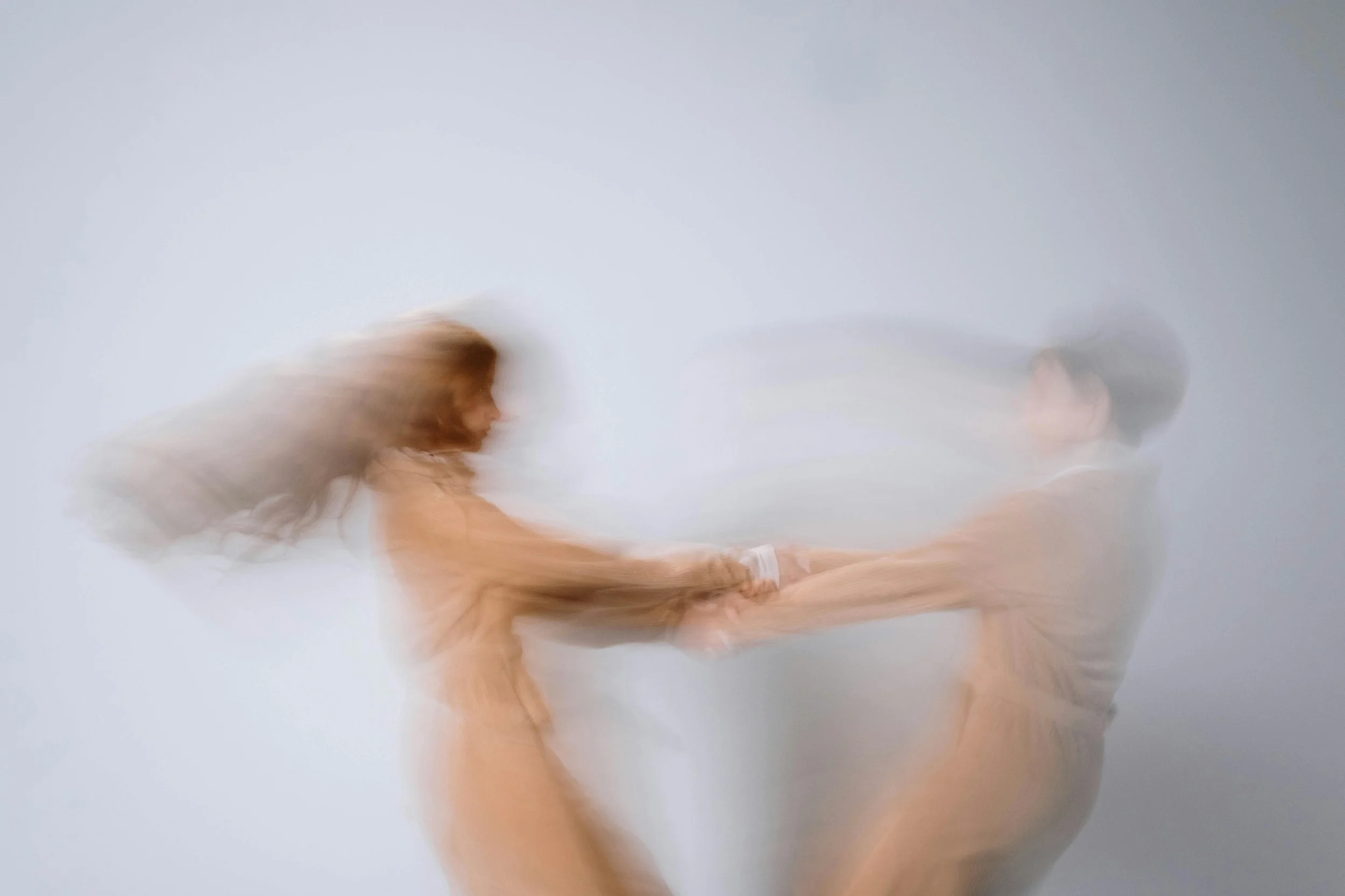 Blurred image of a man and woman holding hands, with the woman leaning back and the man pulling her forward against a plain background.