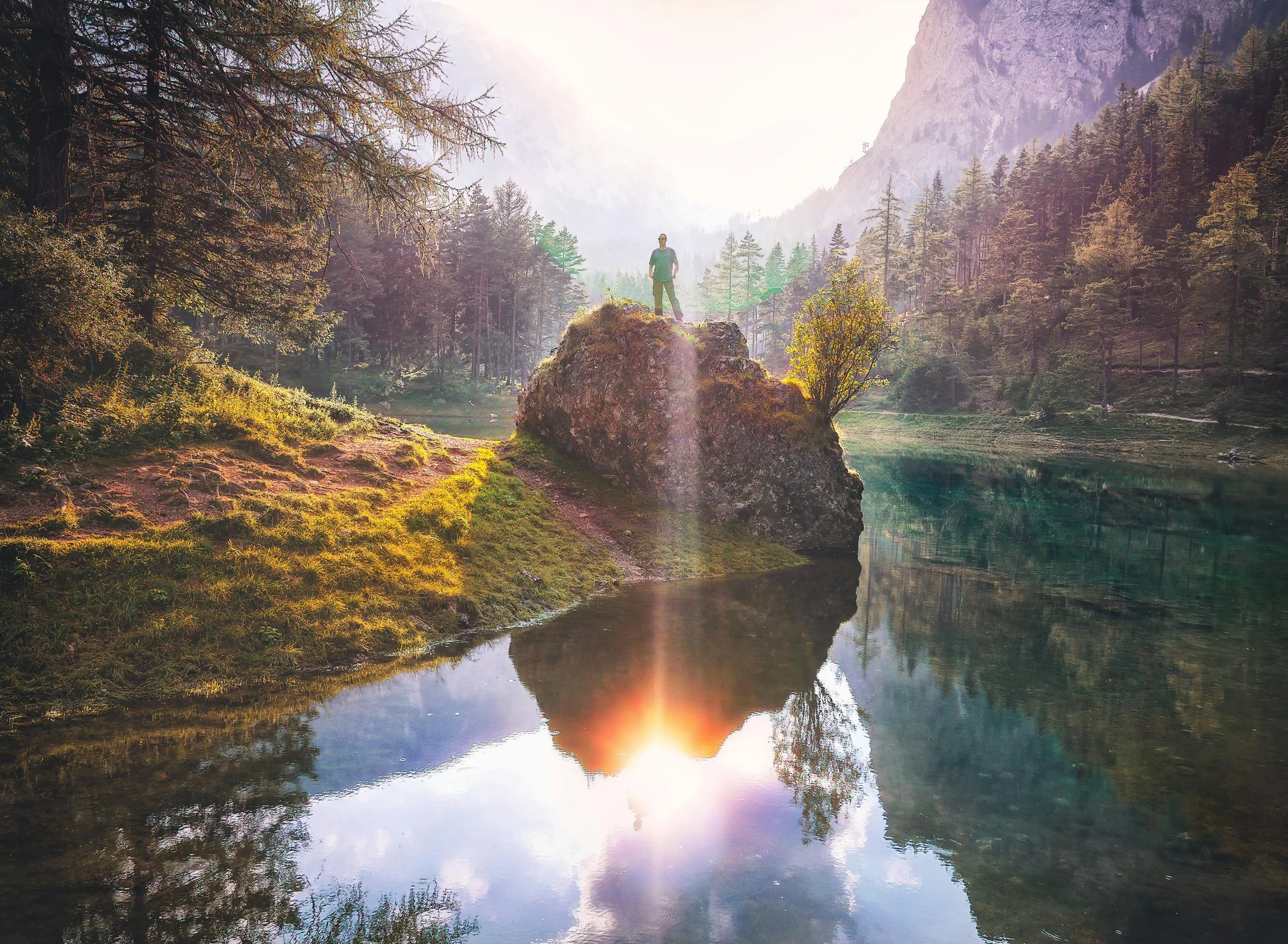 A person standing on a large rock by a tranquil lake in a forested mountainous area during sunrise or sunset, with trees and mountains reflected in the water.