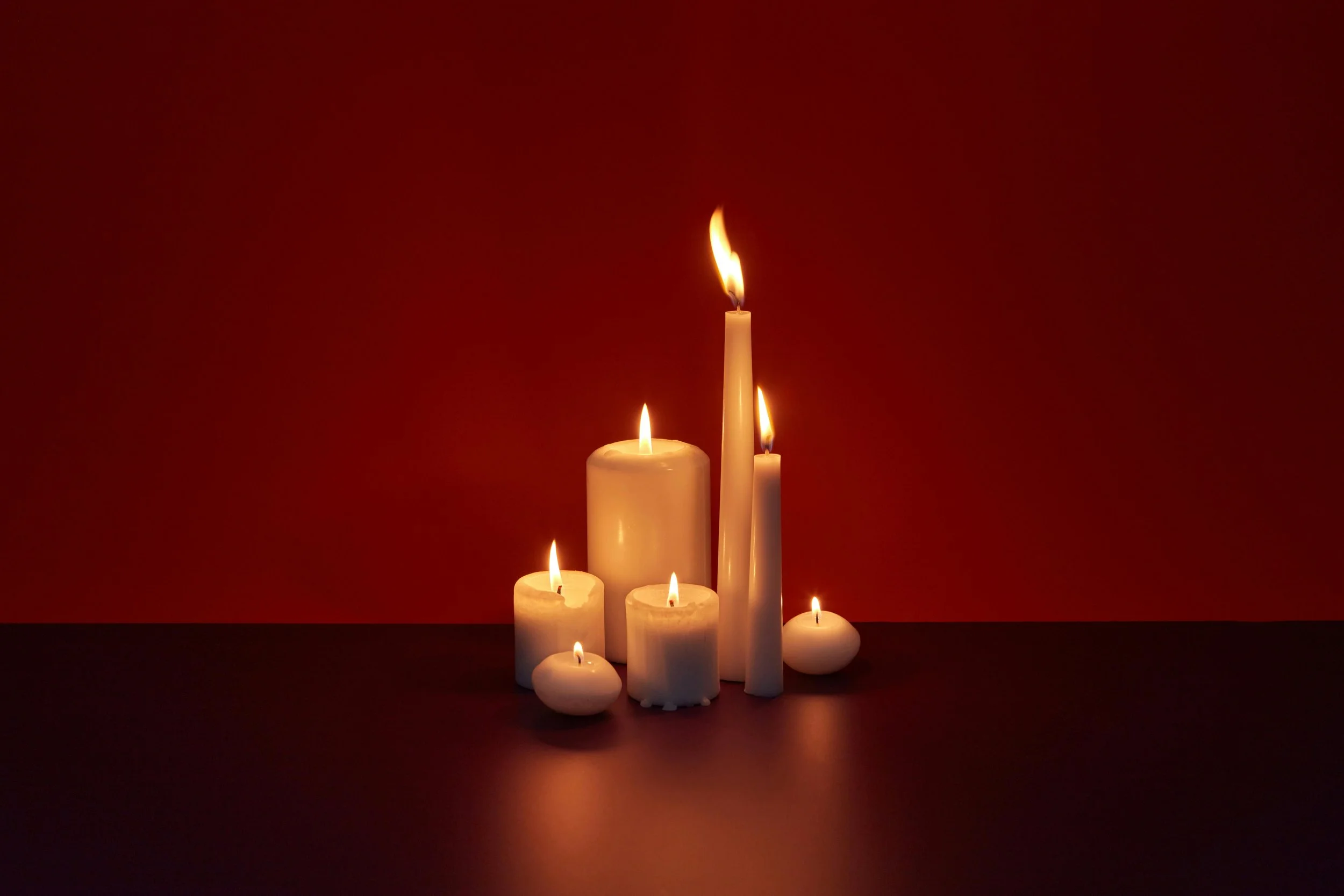 Six lit candles of various sizes on a dark surface against a red background.