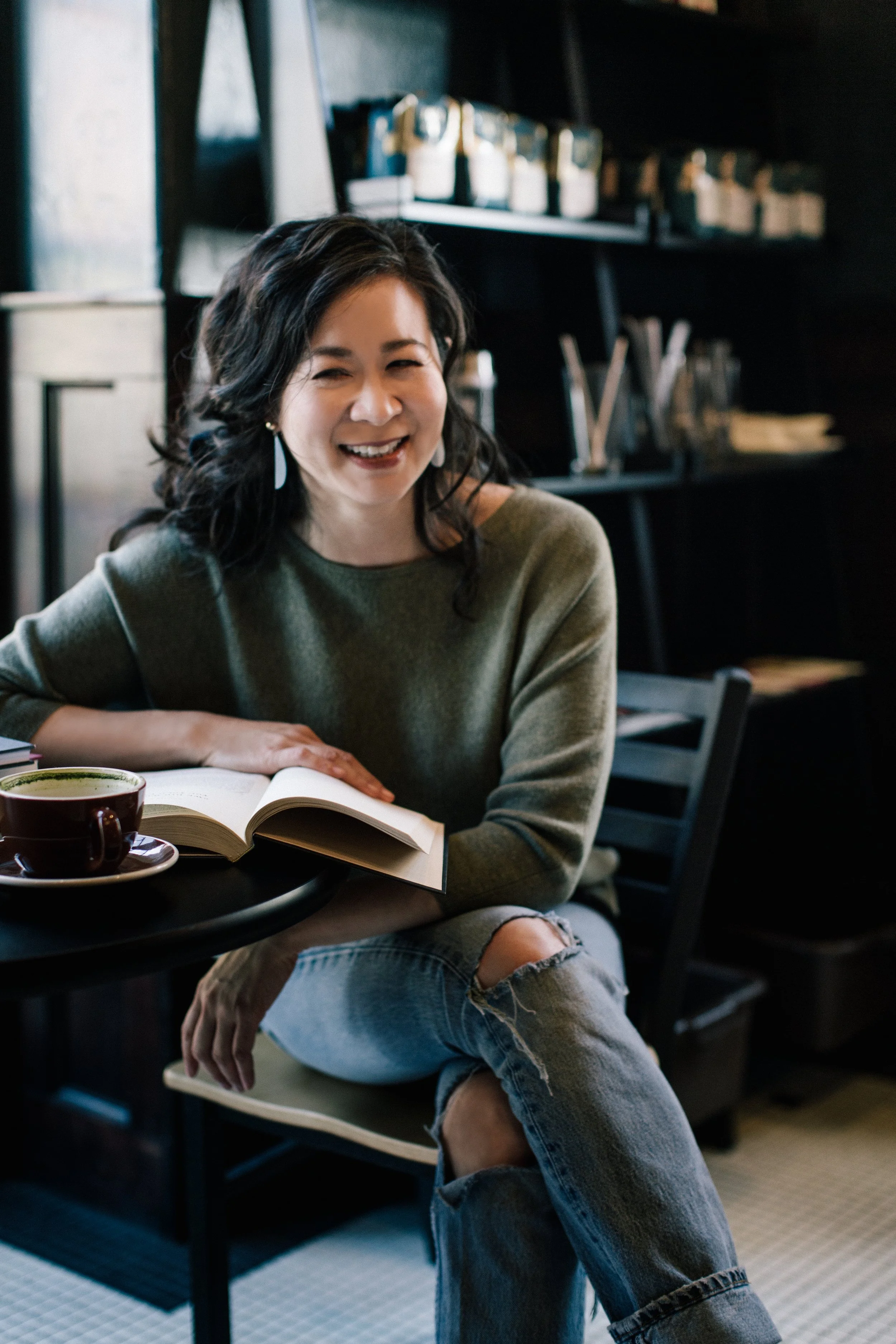 Cindy Kay with dark, wavy hair smiling and sitting at a table in a cozy coffee shop, with an open book and a cup of coffee in front of her.