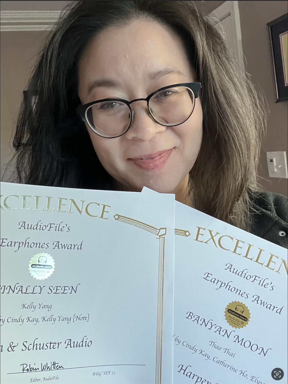 Cindy Kay with glasses and long dark hair holding two awards from AudioFile, smiling.