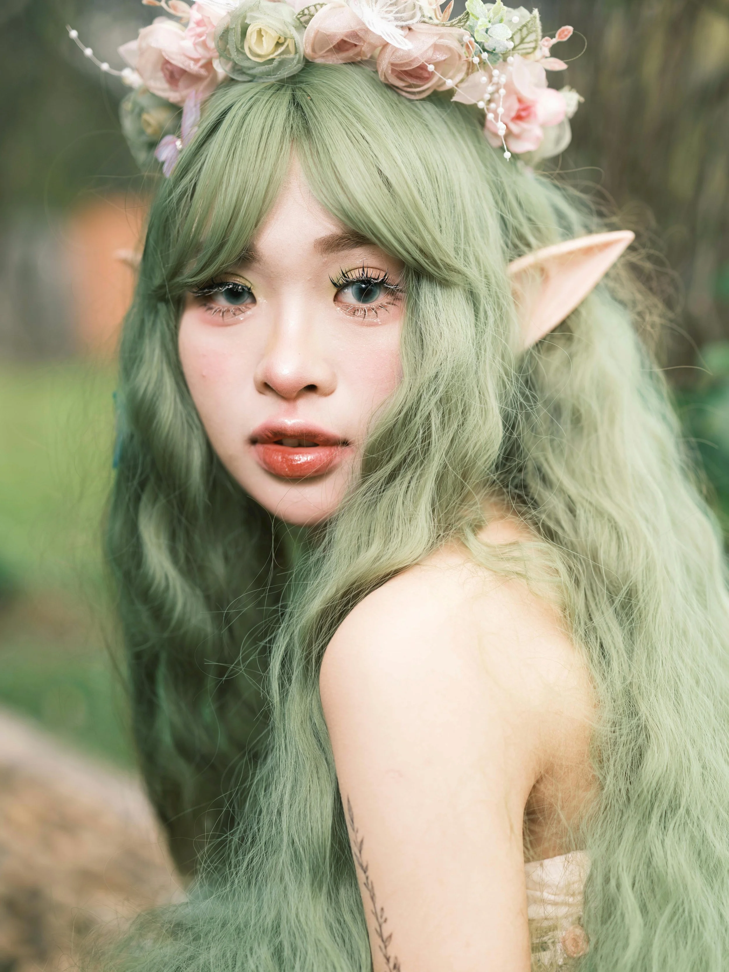 A woman with long, wavy green hair and elf-like ears wearing a flower crown, outdoors. She has makeup with full lips and large eyes, and tattoos on her arm.