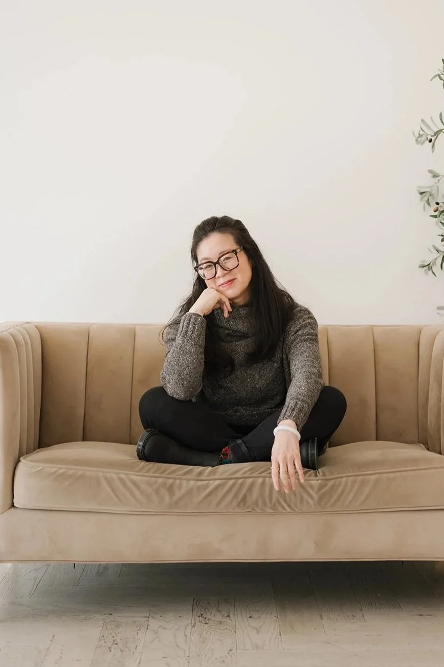 Cindy Kay with long dark hair and glasses sitting cross-legged on a beige vintage sofa against a plain white wall, with her head resting on her hand and smiling.