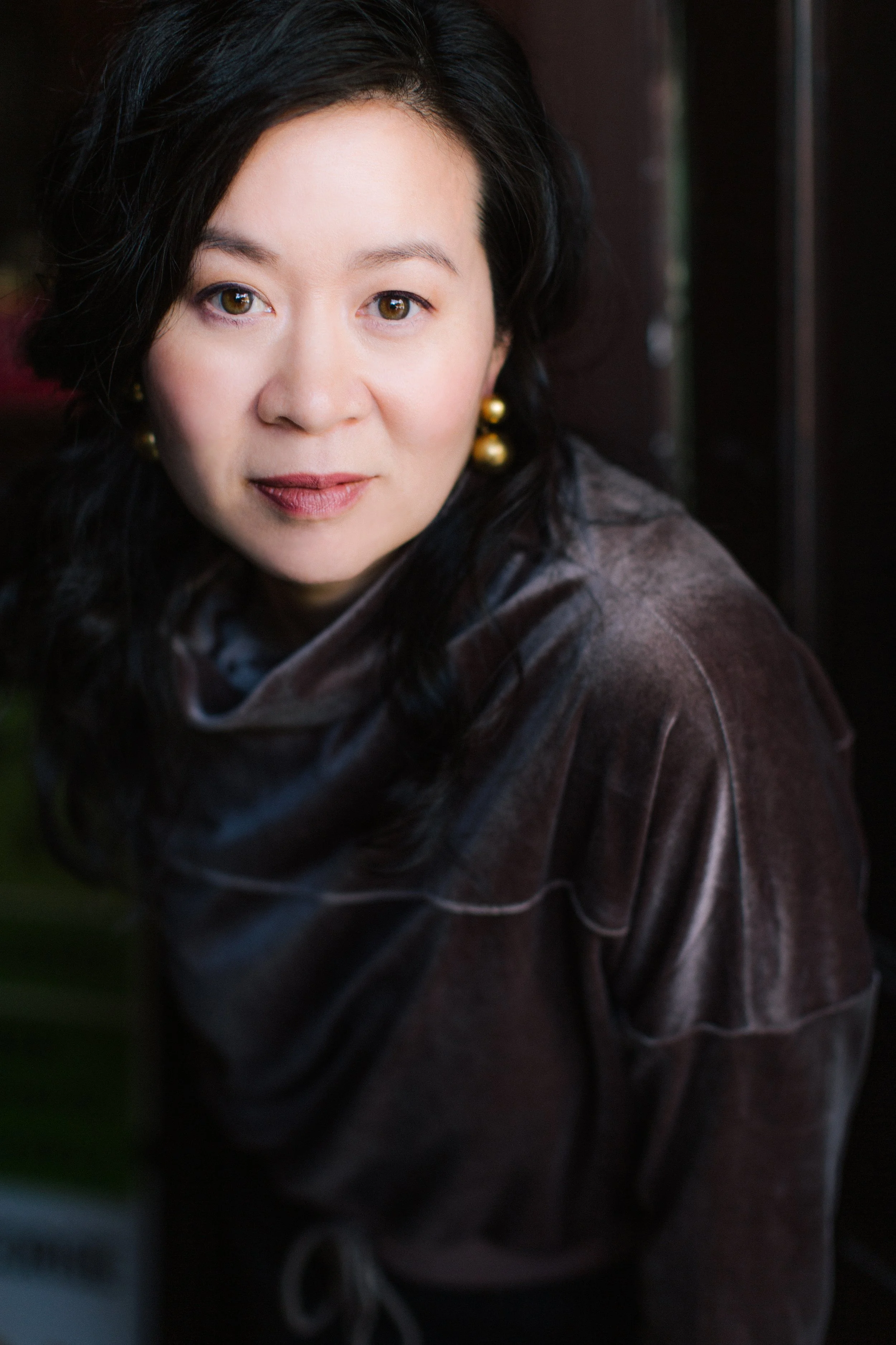 Cindy Kay with black hair and gold earrings looking directly at the camera, wearing a brown leather jacket.
