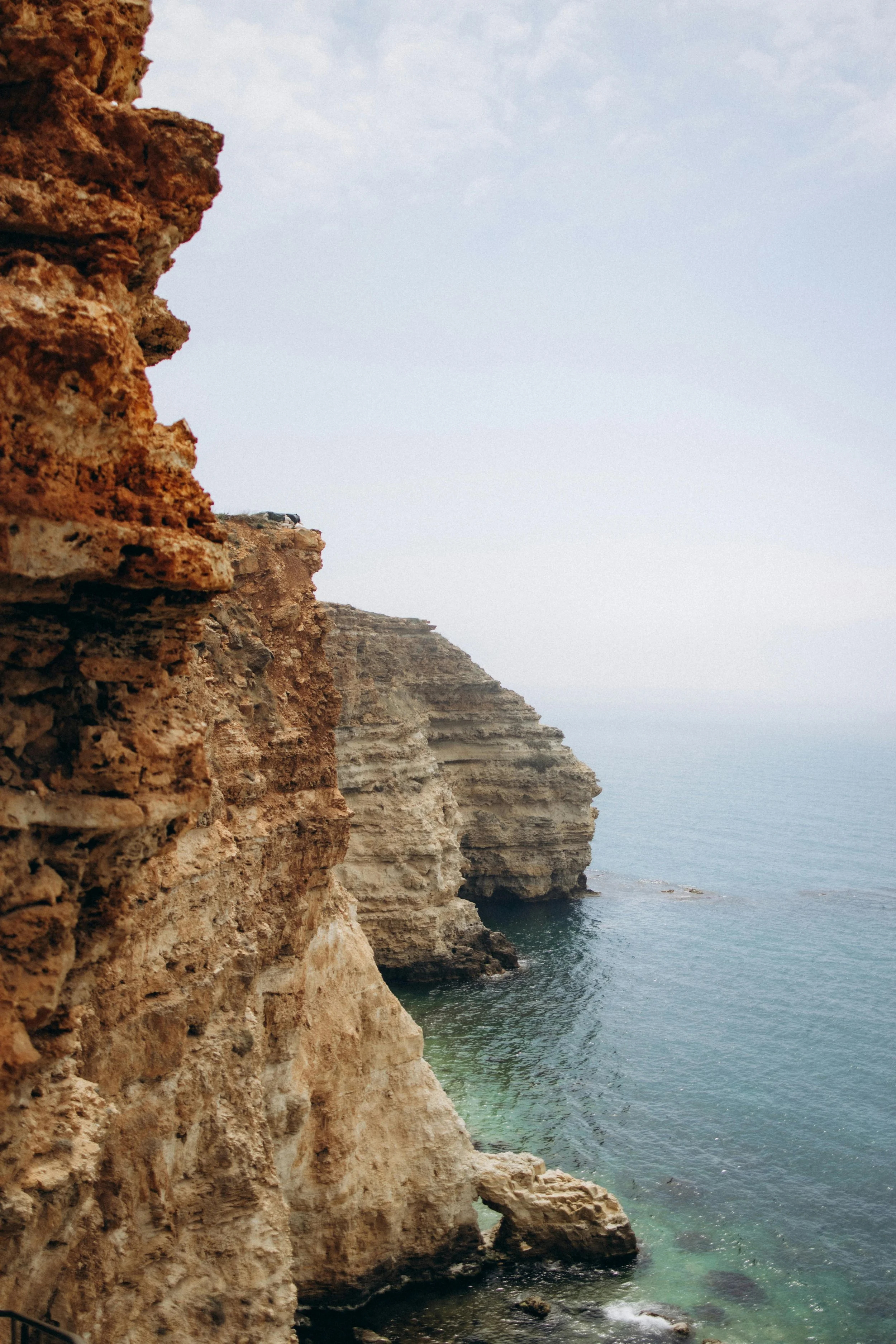 Cliffs overlooking the ocean with a small cave at the base, under a partly cloudy sky.