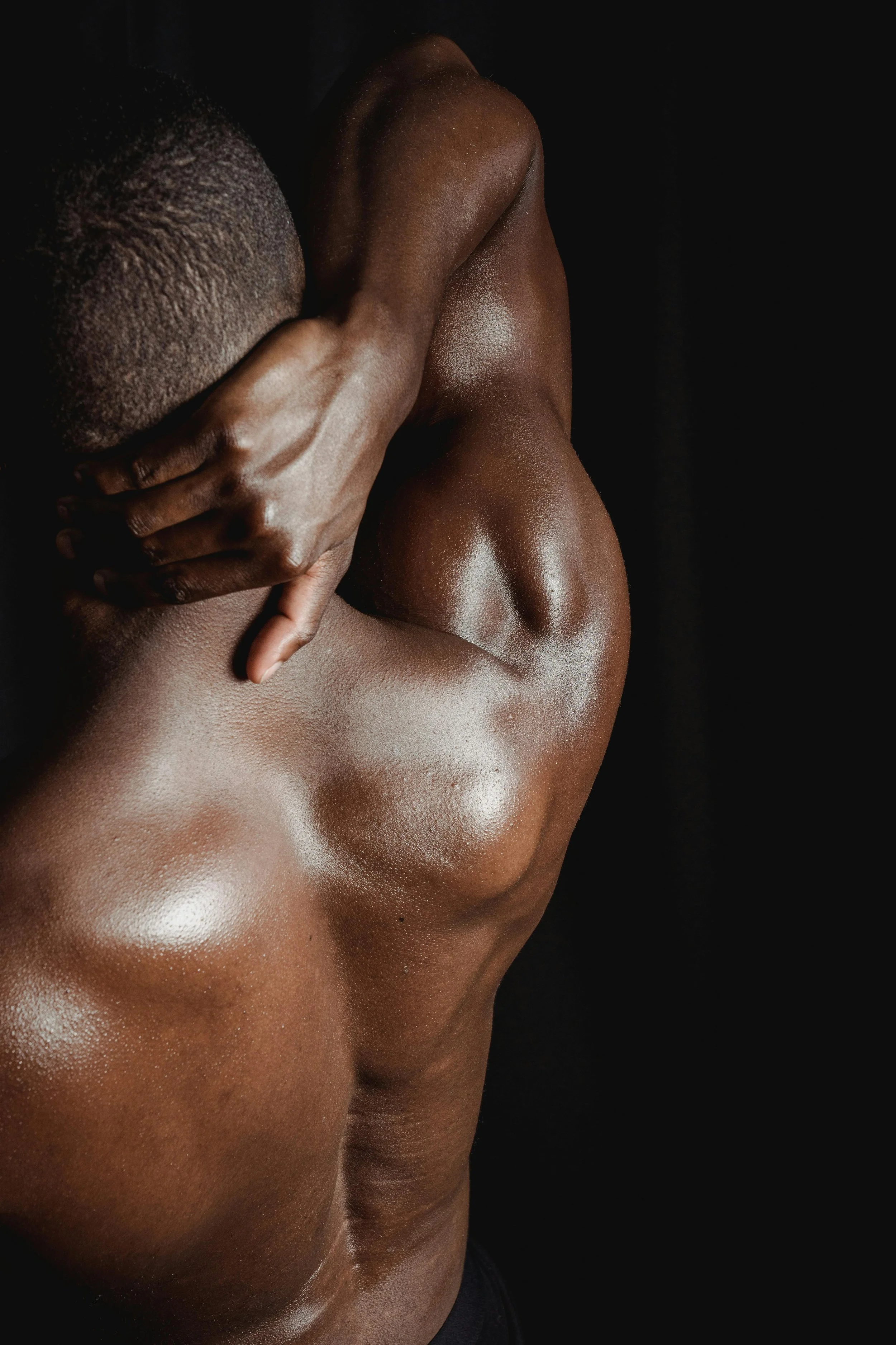 Close-up of a shirtless person with dark skin, holding their neck with one hand, against a dark background.