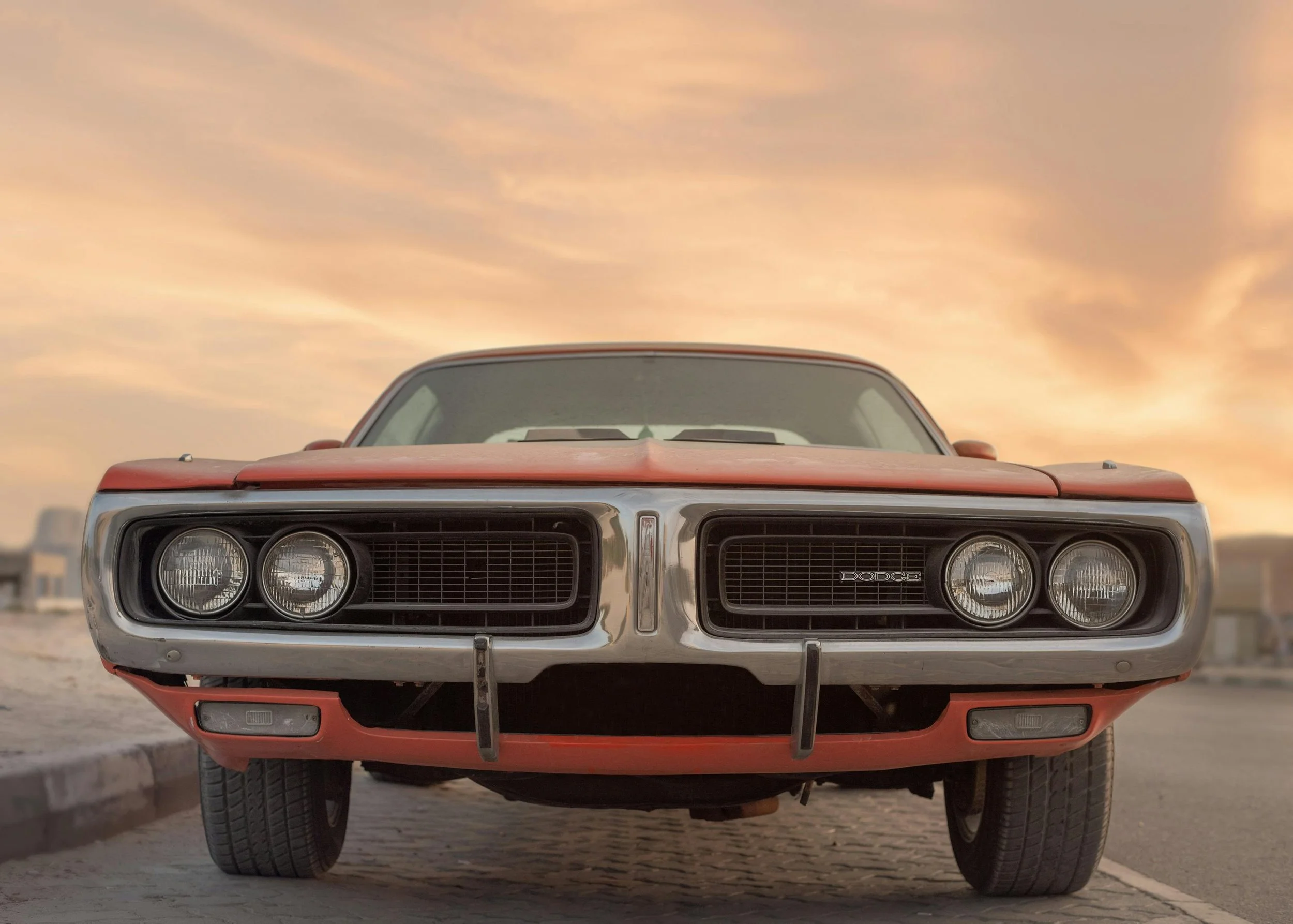 Front view of a vintage Dodge muscle car parked on the street during sunset with a partly cloudy sky.