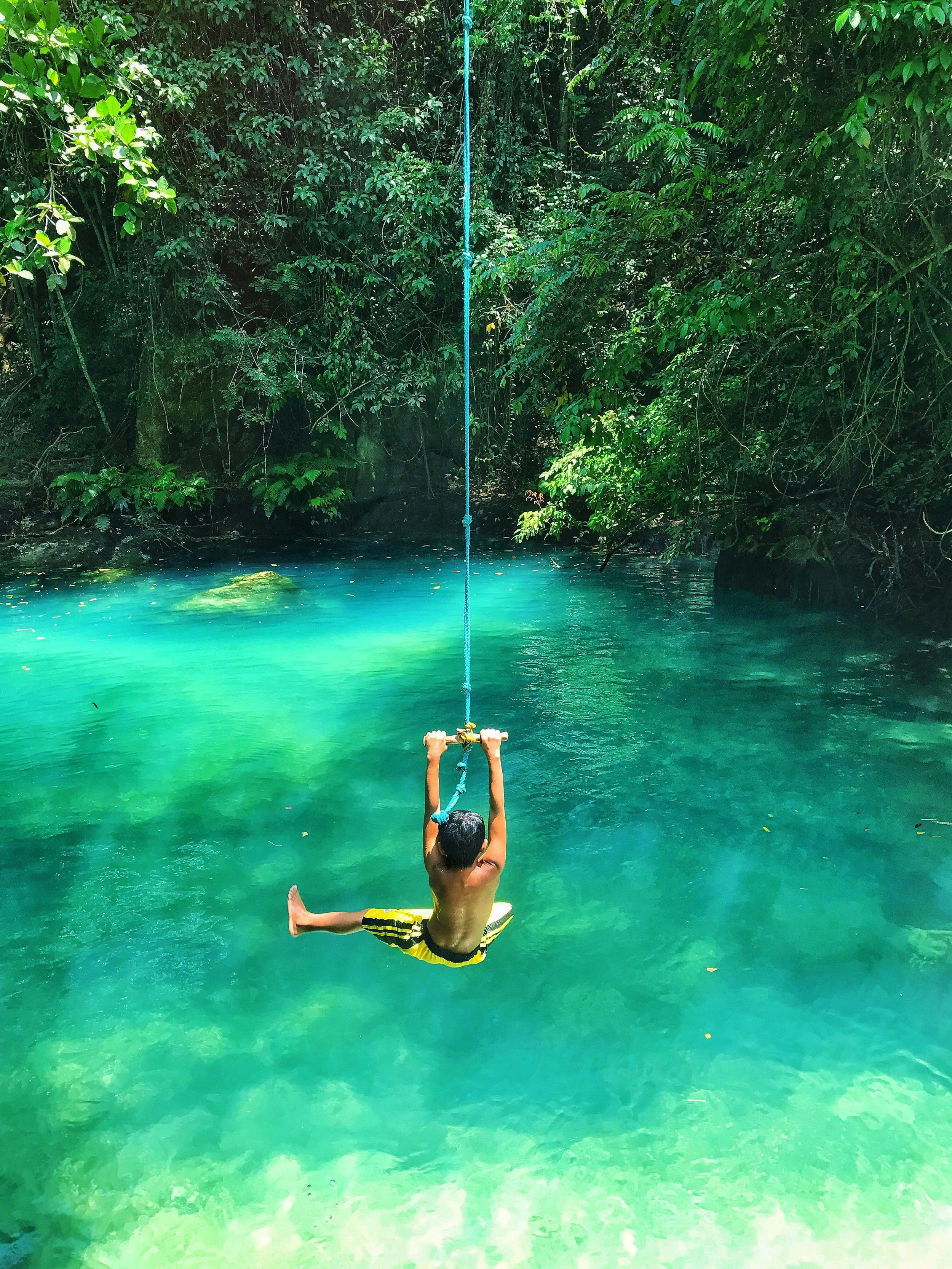 A young boy swinging on a blue rope over a clear turquoise body of water surrounded by lush green foliage.