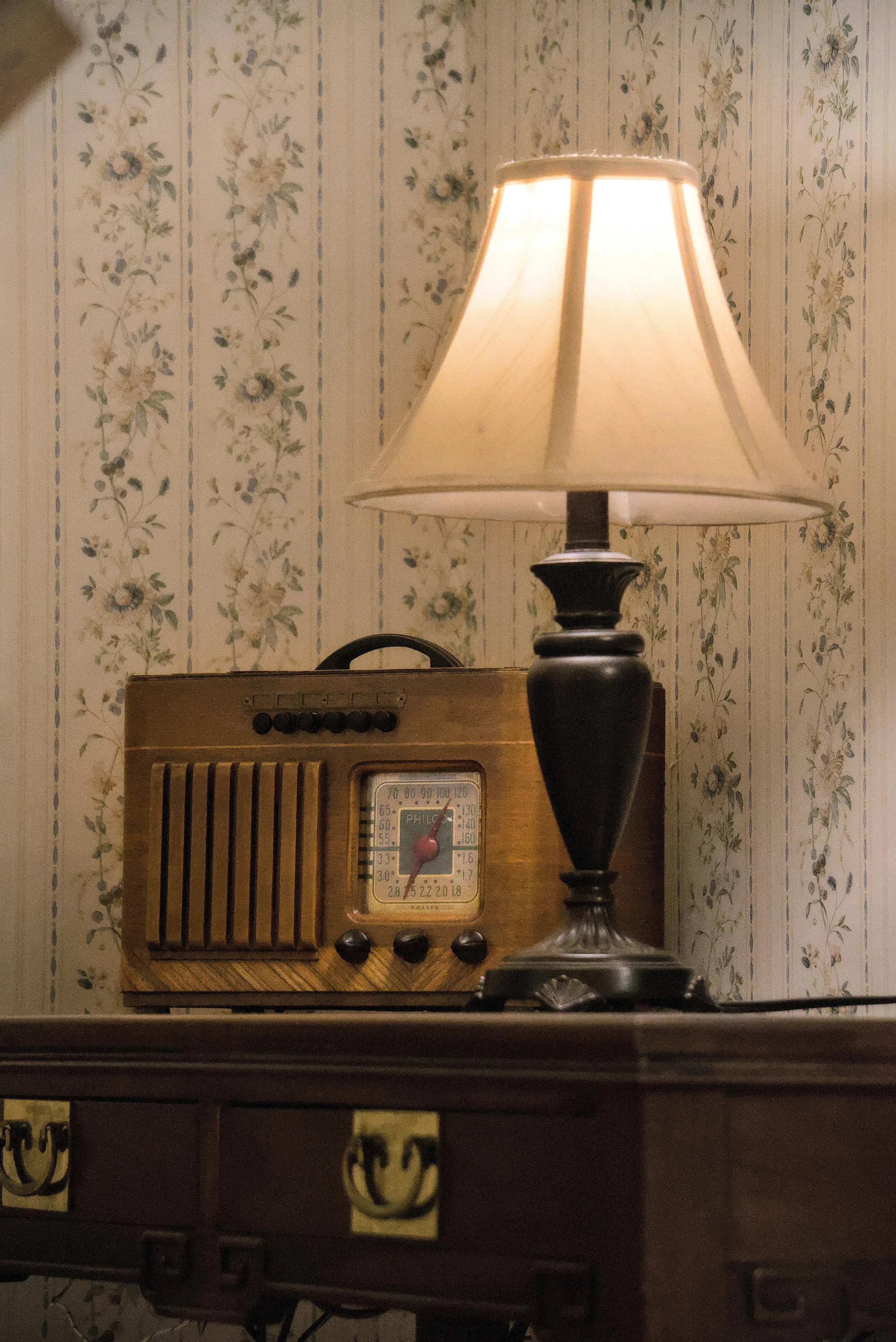 A vintage wooden radio and a table lamp with a beige lampshade on a dark wooden table against floral wallpaper.
