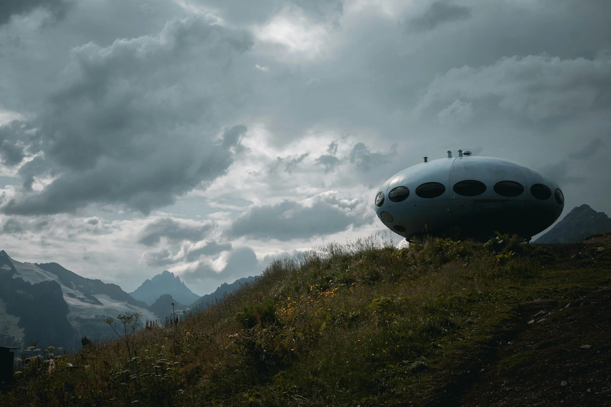 A futuristic, UFO-shaped building sits on a grassy hillside amidst mountains and a cloudy sky.