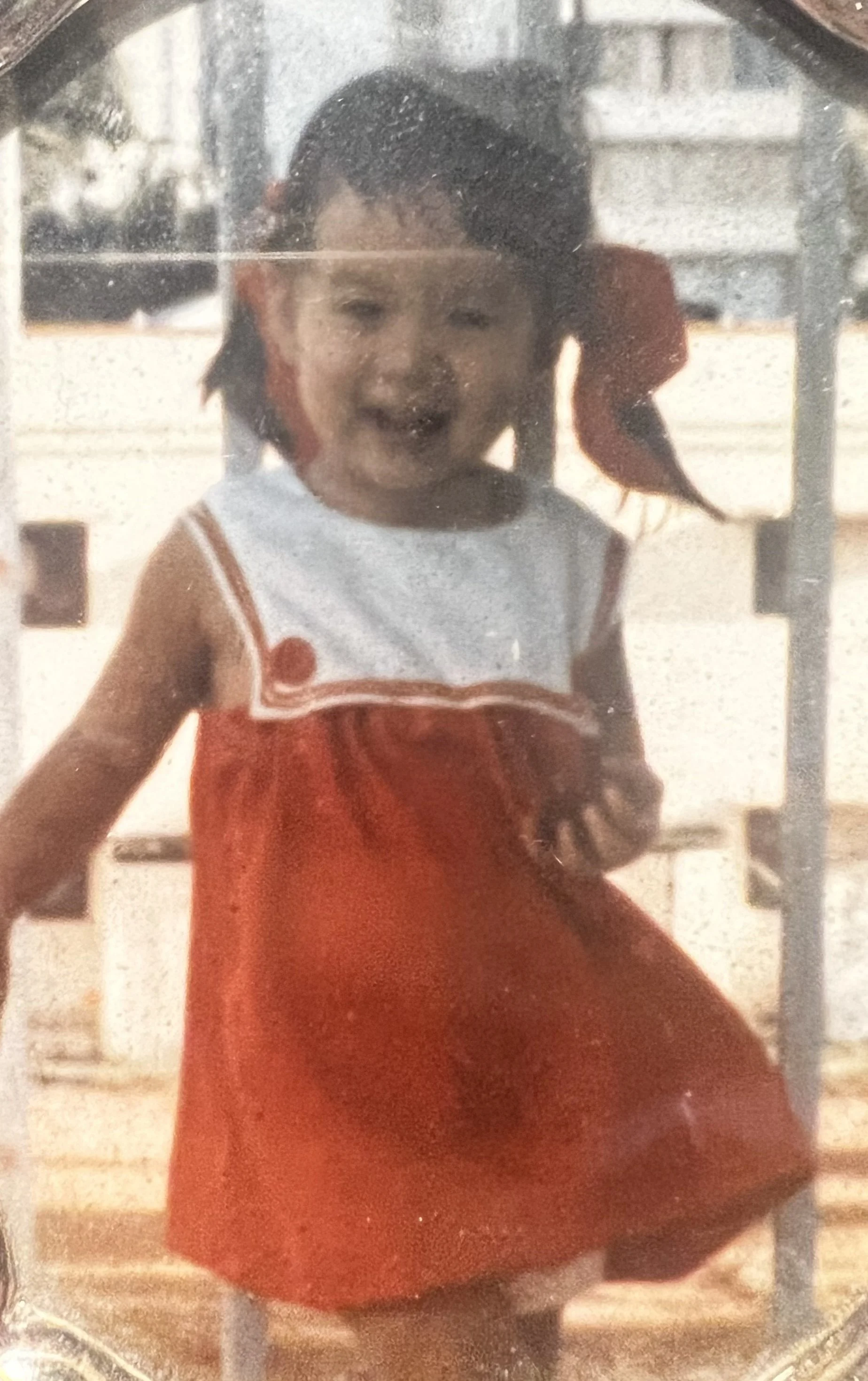 A young girl with pigtails smiling on a playground, wearing a red dress with a white top.