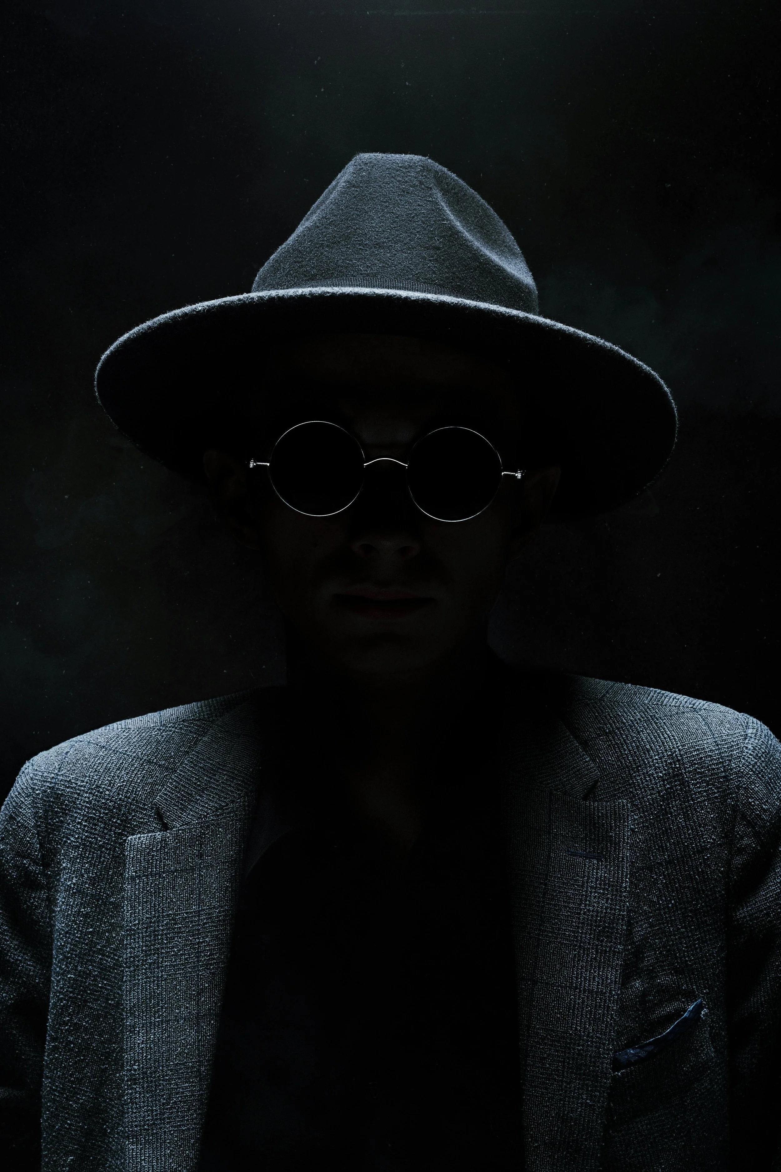 Person wearing a hat and round sunglasses in a dark setting with minimal lighting.