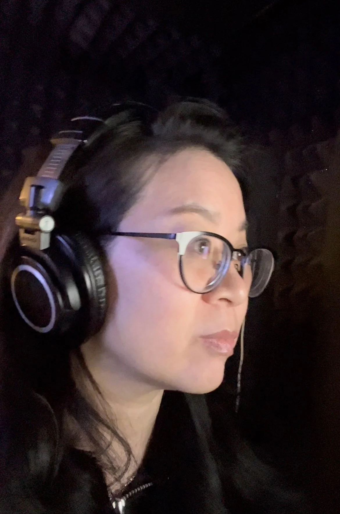 Cindy Kay wearing glasses and headphones in a recording studio, with soundproofing foam on the walls.