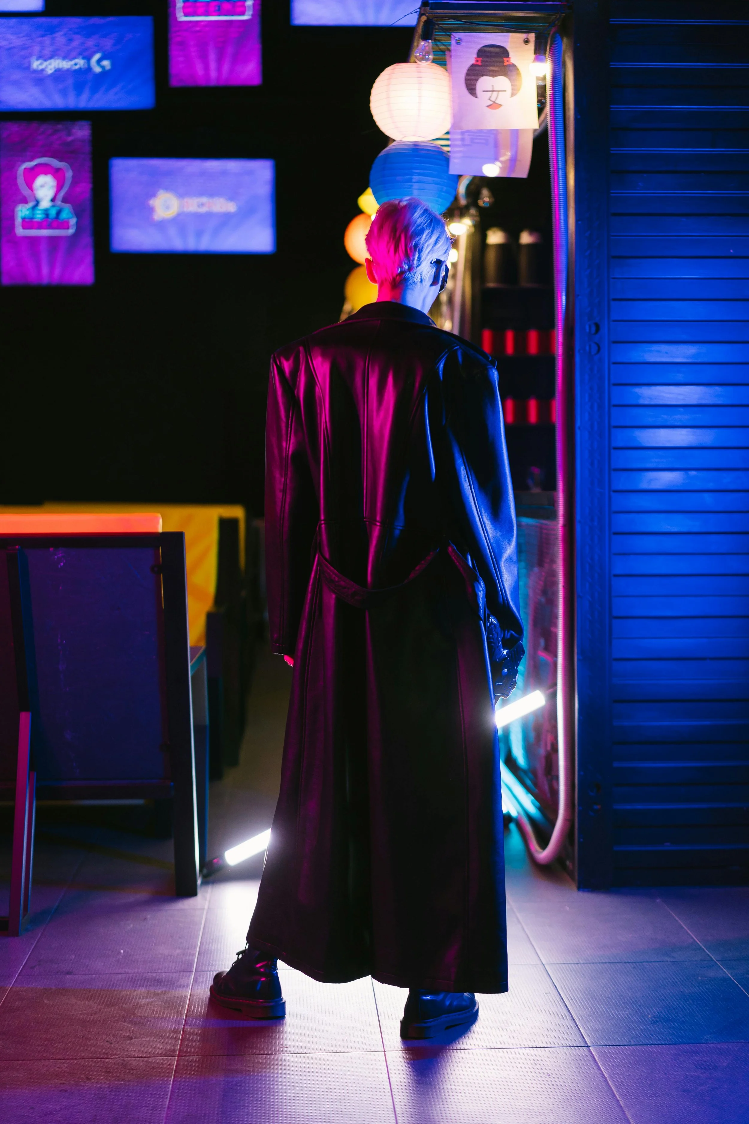 Person in a black leather trench coat and boots standing in an arcade with neon lights; rear view.