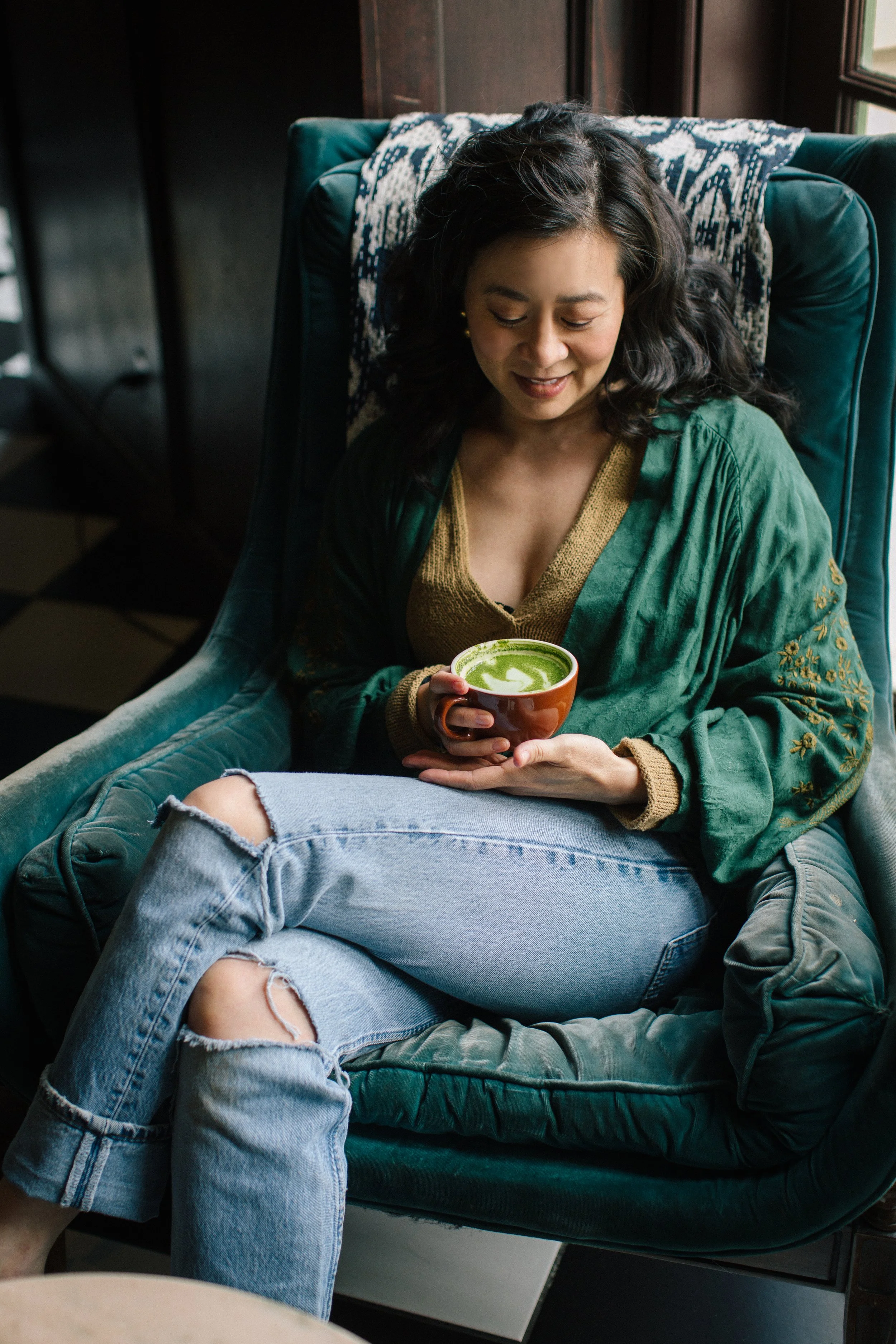 Cindy Kay sitting in a green armchair, holding a green latte in a mug, in a cozy indoor setting near a window.