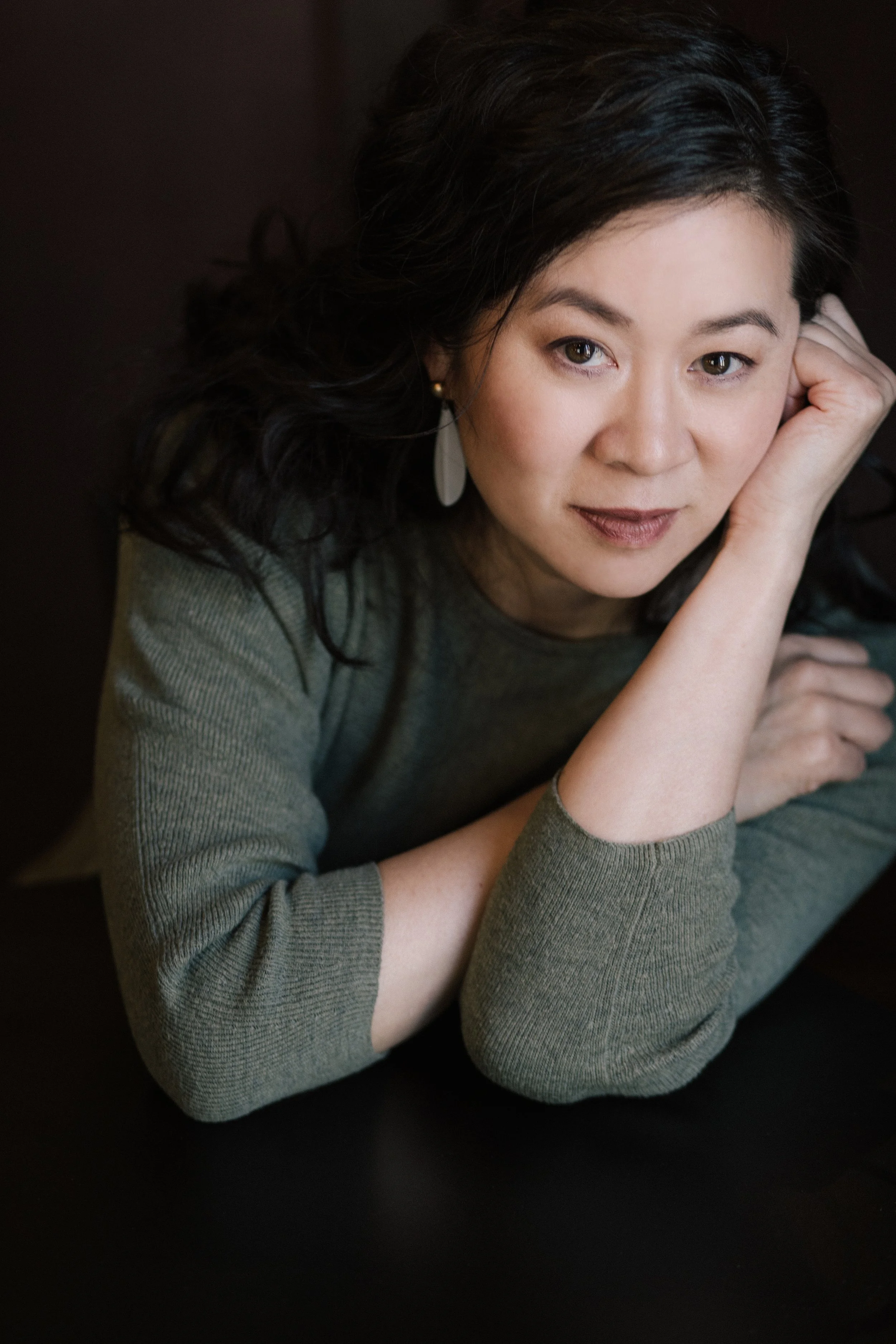 Close-up of Cindy Kay with dark hair, lying on her side with her head resting on her hand, wearing a green long-sleeved top, looking at the camera with a slight smile.