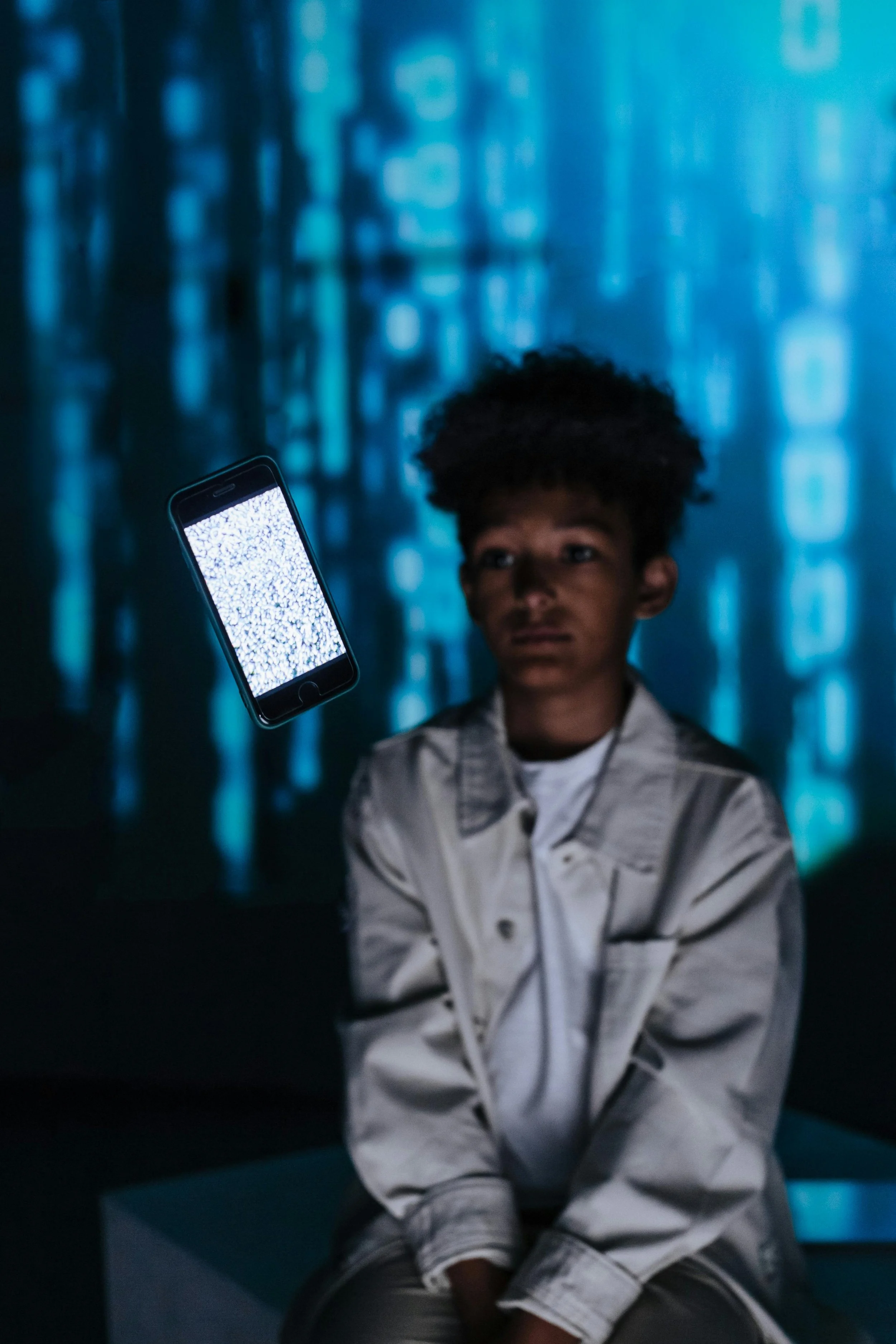 A young person with curly hair sitting indoors, looking at a floating smartphone displaying static. The background is illuminated with blue digital code patterns.