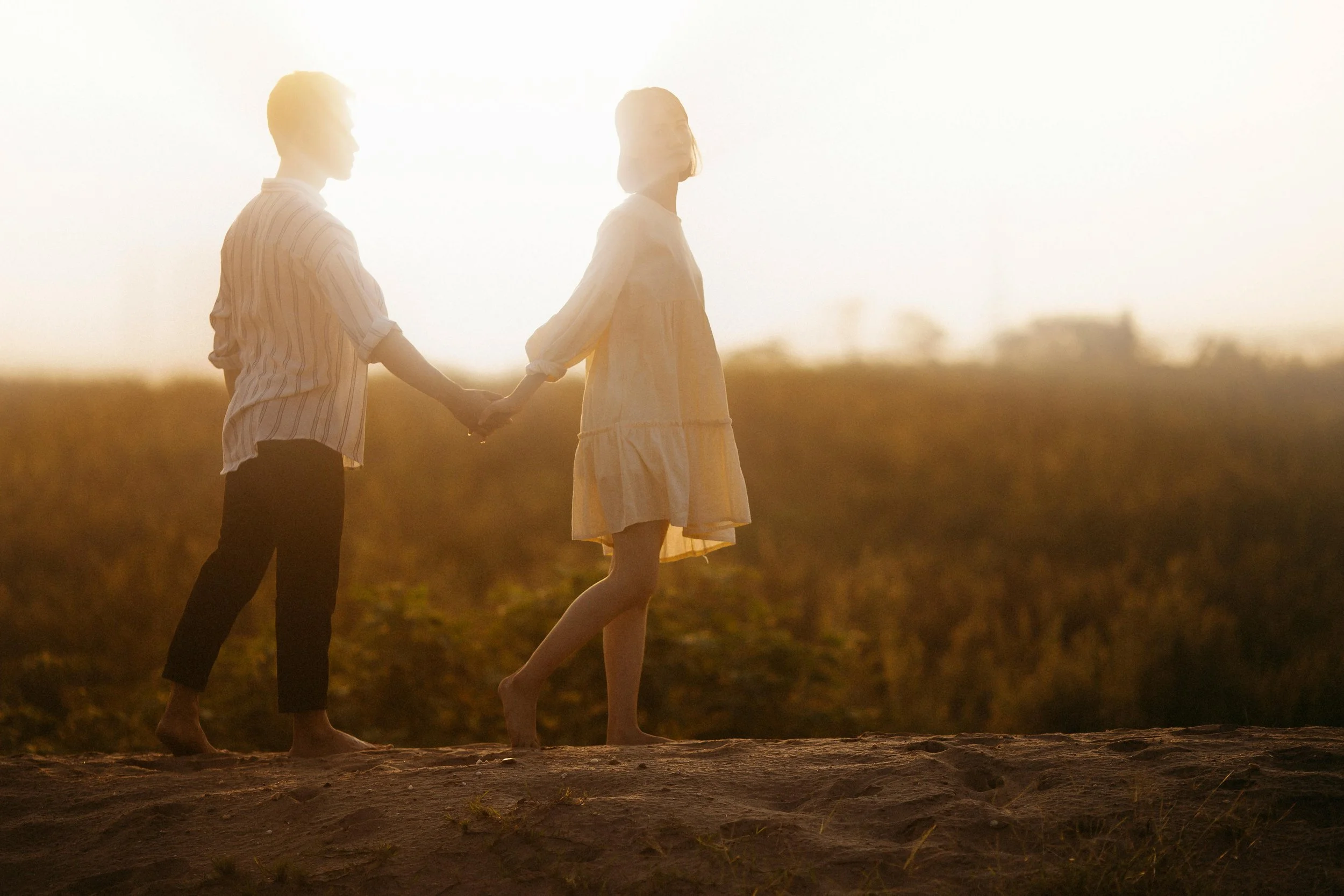 A man and woman holding hands and walking on a dirt path during sunset.