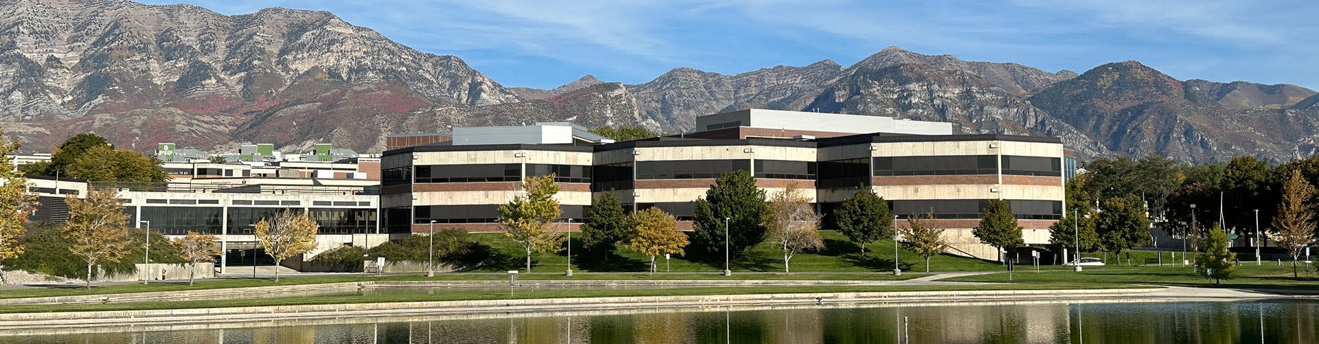 A modern office building with multiple floors, surrounded by trees with fall foliage, beside a water body with mountains in the background under a clear blue sky.
