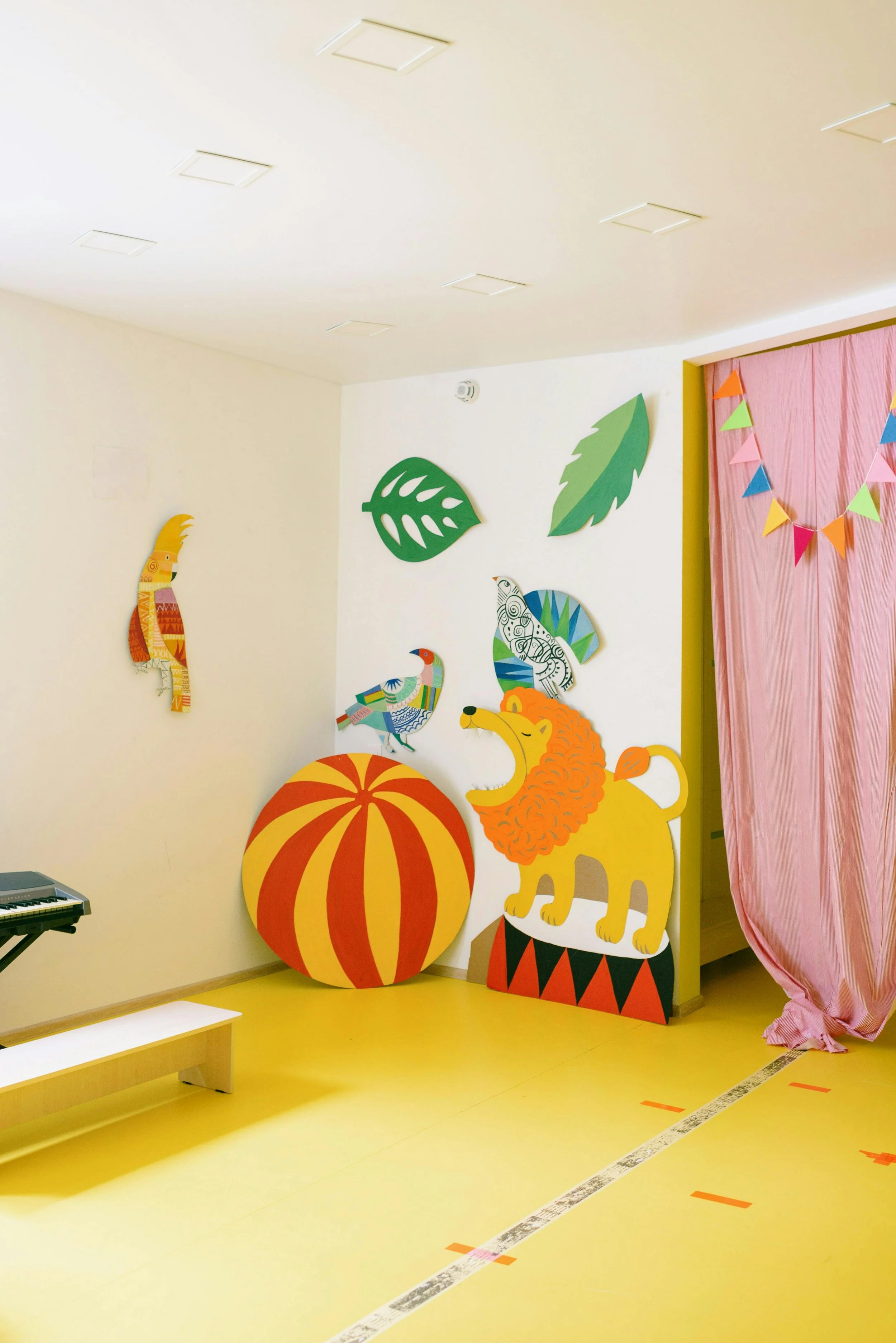 Decorative classroom corner with colorful cardboard cutouts including a lion with a mane, a large orange and yellow pumpkin, four stylized birds, and green leaves. A pink curtain with a multi-colored bunting is on the right side, and a digital keyboard is partially visible on the left.