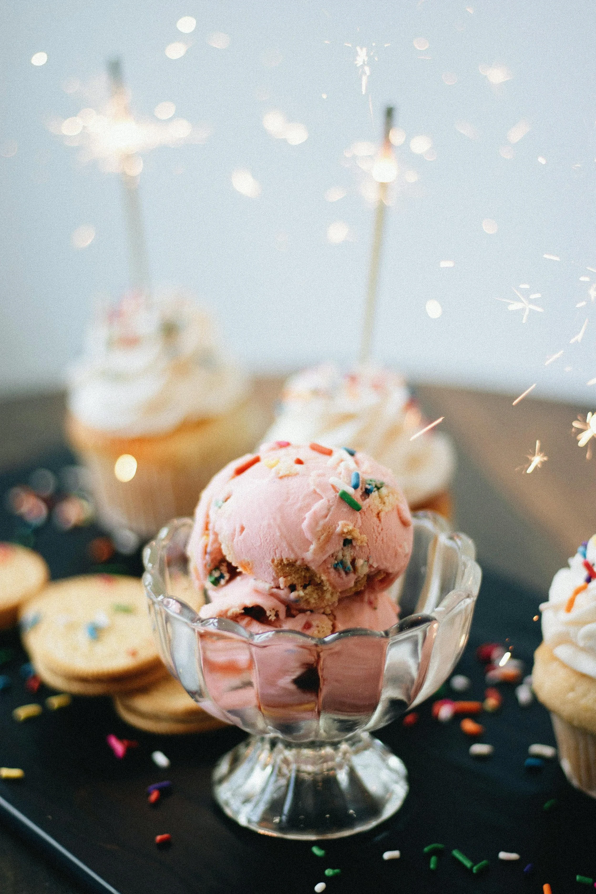 Pink ice cream with colorful sprinkles in a glass dish, surrounded by cookies and cupcakes with whipped cream and sparklers in the background.