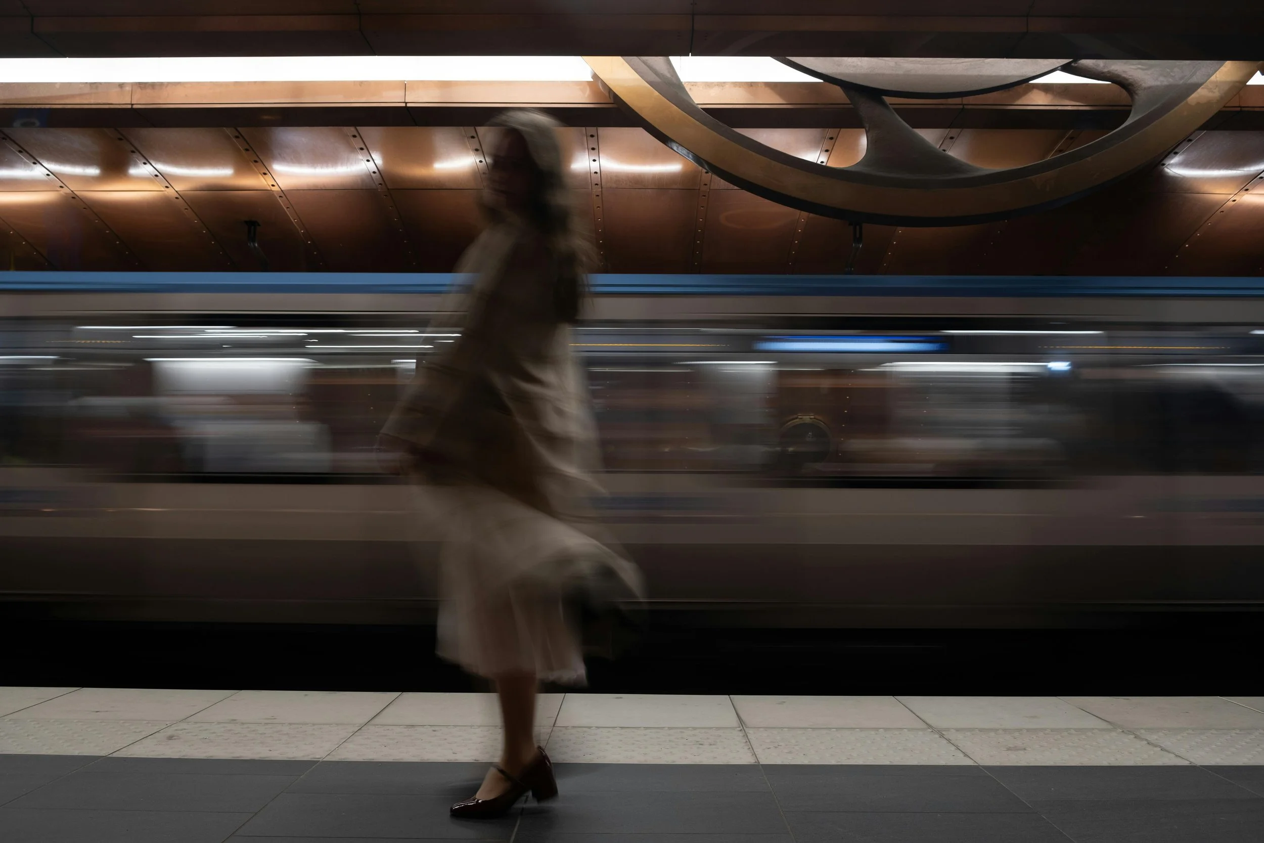Blurred image of a woman in a white skirt and dark shoes walking past a moving train at a subway station with wooden ceiling panels.