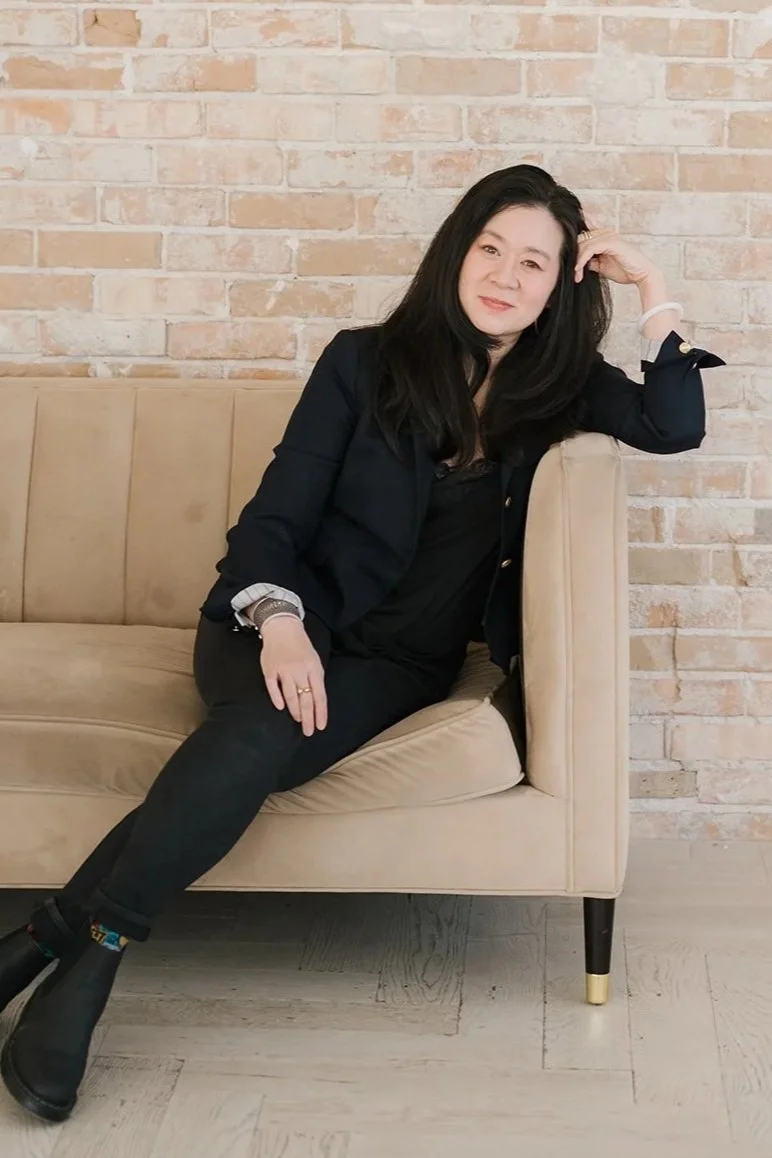 Cindy Kay with long black hair sitting on a beige sofa, resting her arm on the backrest, in front of a brick wall.