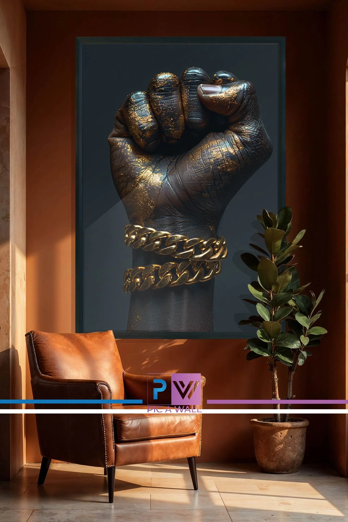A large framed artwork of a black hand with gold paint splatters, wearing two thick gold chain bracelets, hangs on a brown wall. In front of the artwork is a brown leather armchair with black legs, next to a potted green plant with rounded leaves. The scene is lit by natural sunlight.