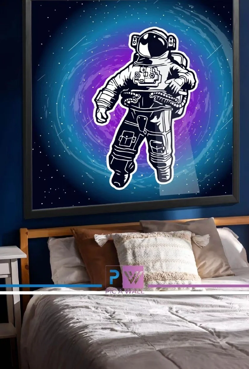 A bedroom with a large framed space-themed artwork of an astronaut in a spacesuit floating in a galaxy with stars and swirling colors. The bed has white, beige, and brown pillows and a white blanket. The room has dark blue walls and a small white sid
