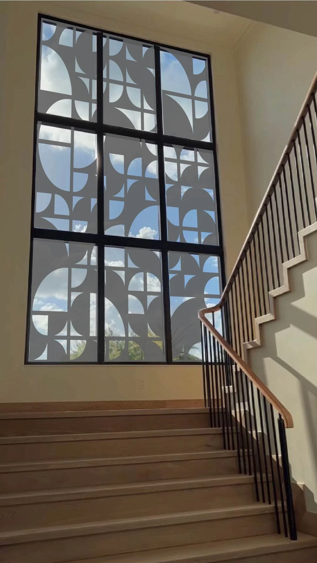 Indoor staircase with wooden steps and black metal railing leading up to a large window. The window has decorative frosted film with abstract circular patterns, letting in sunlight and showing a blue sky with clouds outside.