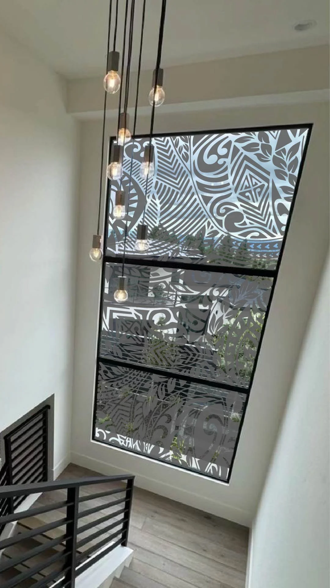 Interior view of a modern staircase with black railing, a large window with decorative patterned glass, and hanging pendant lights.