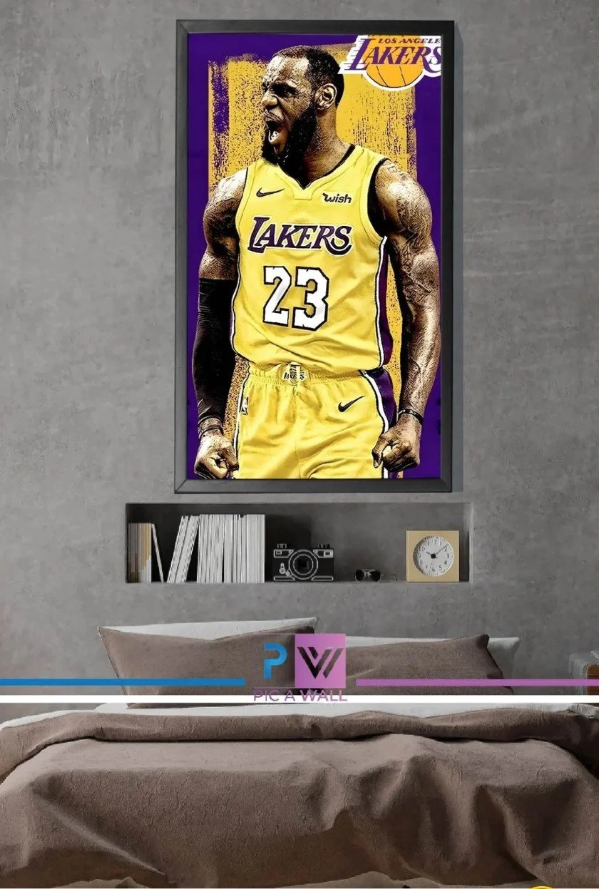 A framed poster of LeBron James in a Los Angeles Lakers uniform with the number 23, celebrating with an angry expression, hanging on a gray wall above a bed. Below the poster is a small shelf with books, a camera, a clock, and small objects.